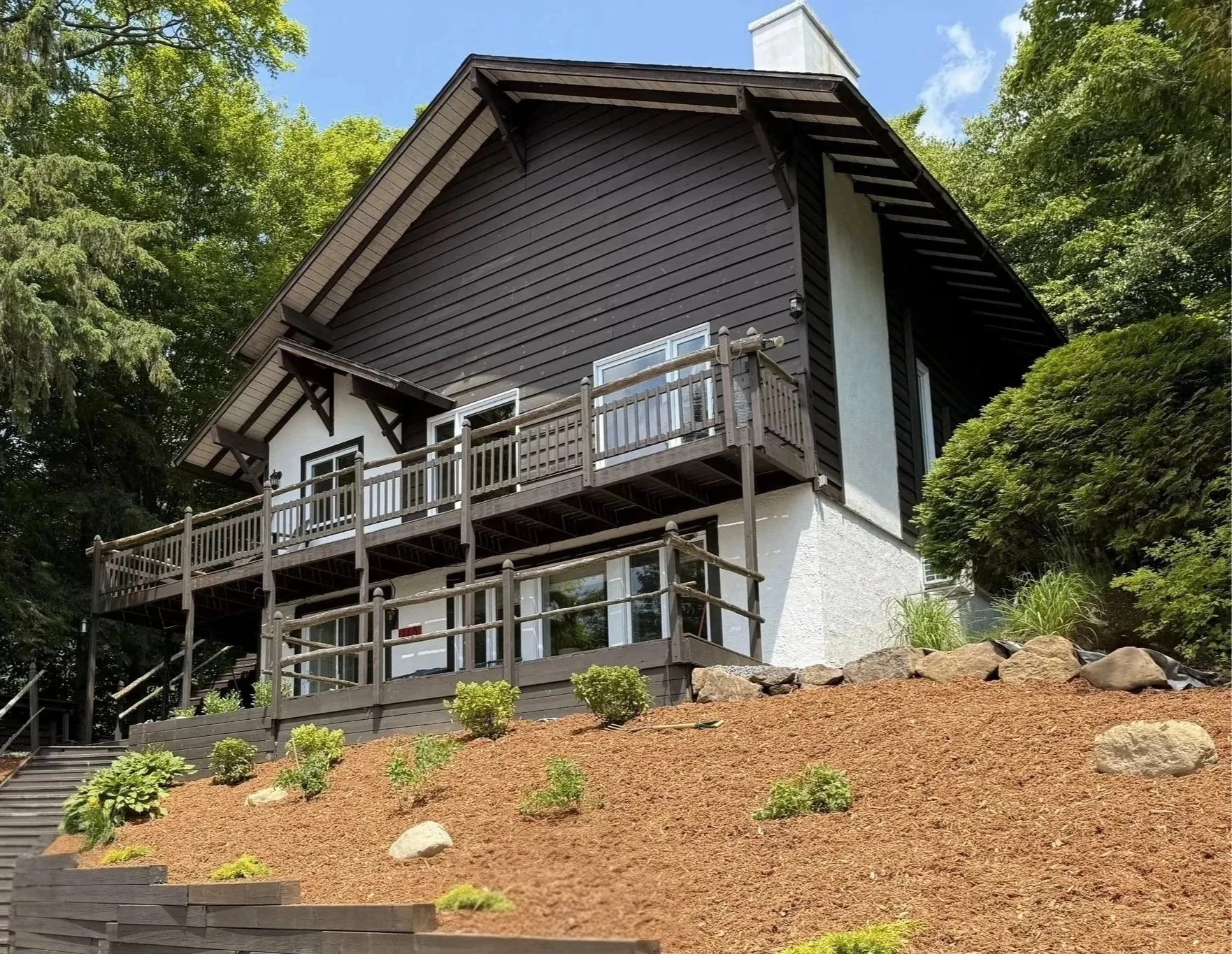 Une maison en bois située sur une pente, entourée d'arbres verts, avec deux niveaux de terrasse en bois, en partie en angle, sous un ciel bleu avec quelques nuages.