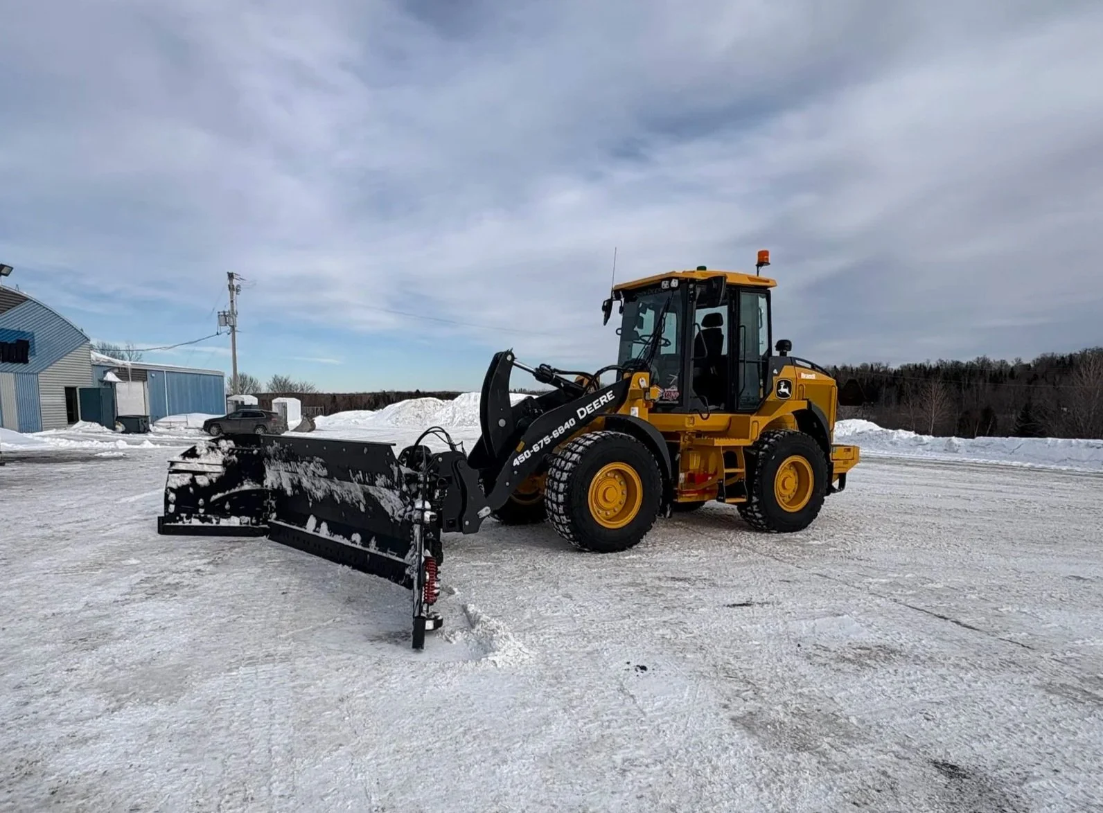 Véhicule de déneigement jaune avec lames en avant, stationné sur une surface enneigée. Le ciel est nuageux, avec quelques bâtiments et une voiture en arrière-plan.