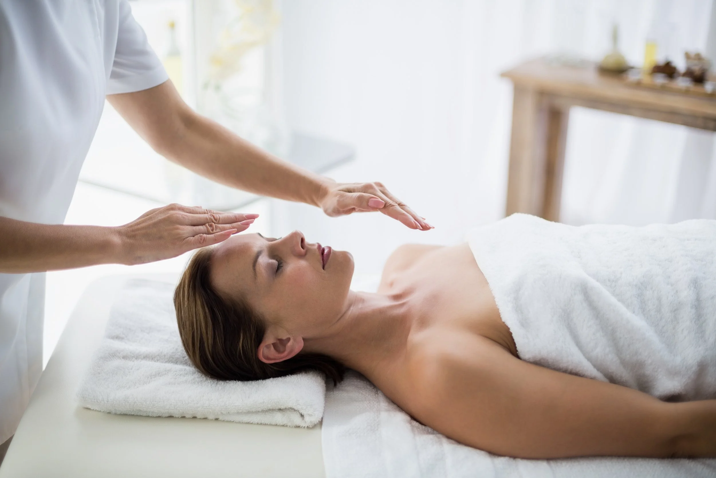 A woman lying on a treatment table with her eyes closed, receiving a facial massage or treatment from a professional in a bright, clean spa or clinic setting.