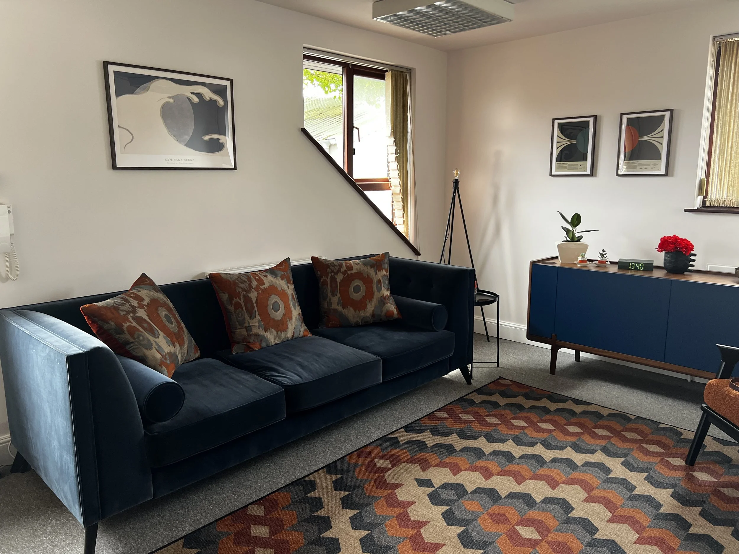 A cozy living room corner with a dark blue sofa adorned with patterned pillows, a colorful geometric area rug, artwork on the white walls, a window with sunlight and curtains, a small table with a plant and a digital clock, and a floor lamp.