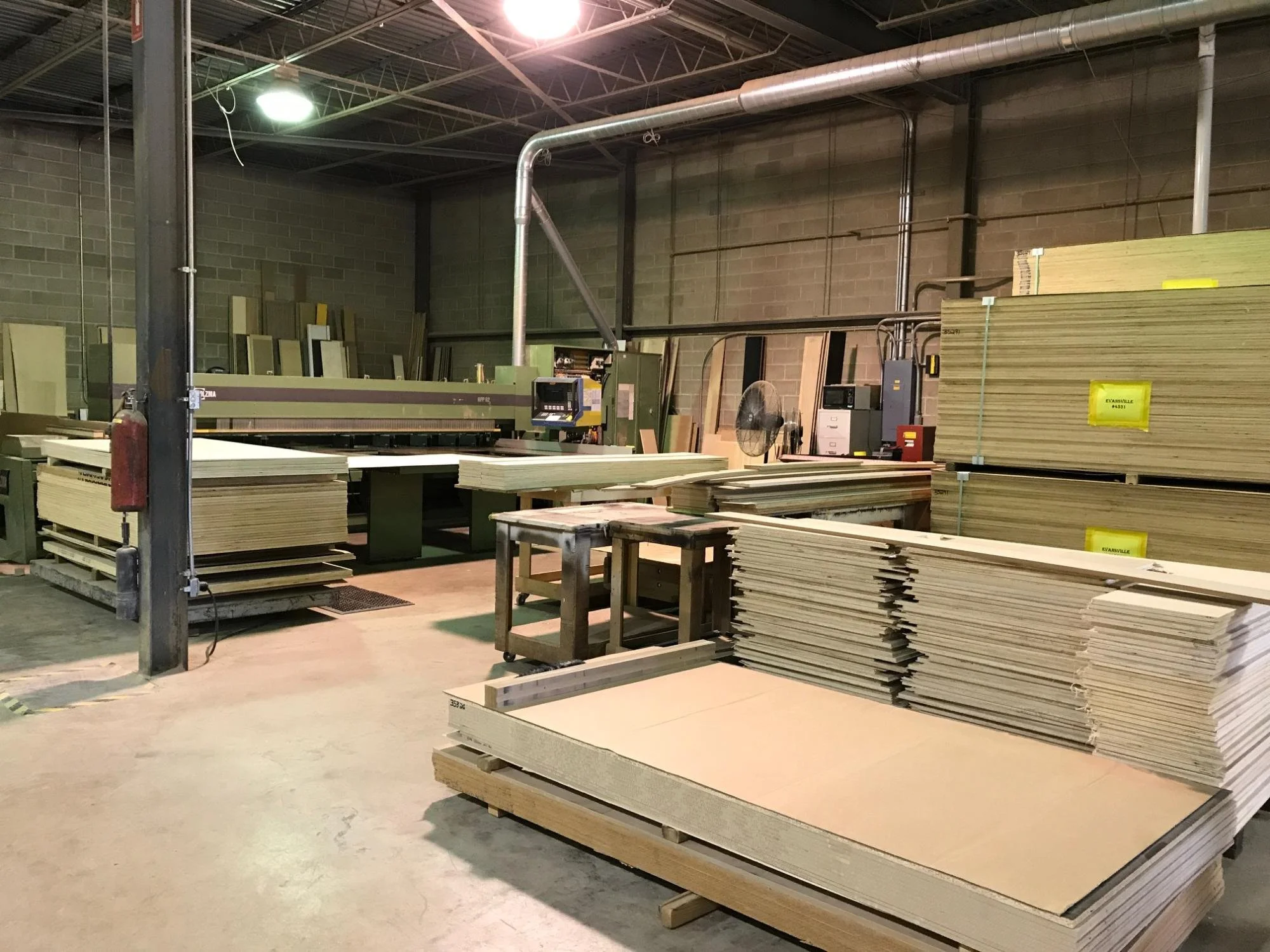 Interior of a woodworking shop with stacks of plywood and lumber, a large cutting machine, and various tools.