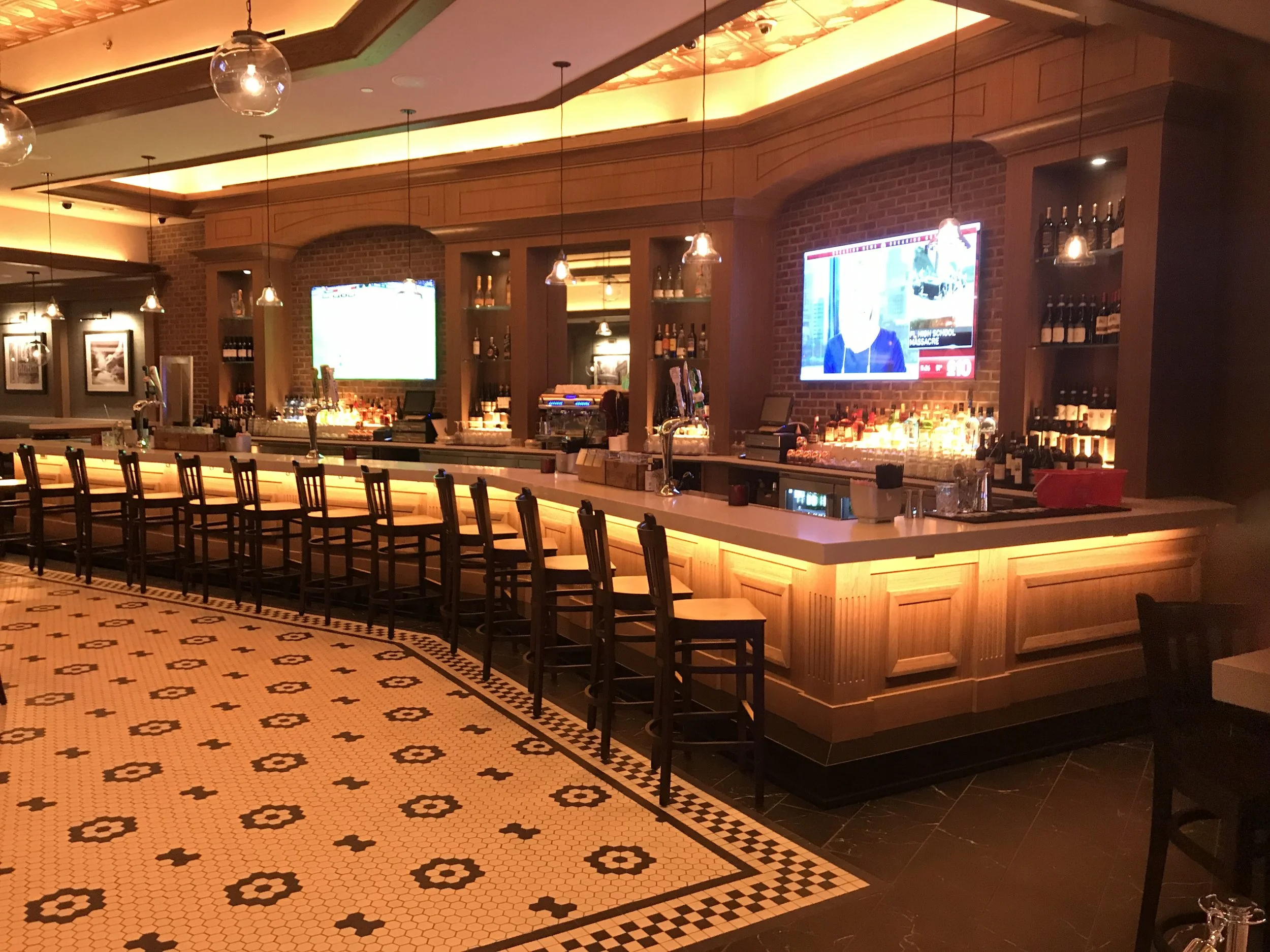 Empty bar with wooden counter and high chairs, illuminated by warm lighting, with TV screens on brick wall, bottles of alcohol on shelves, and black and white patterned tile floor.