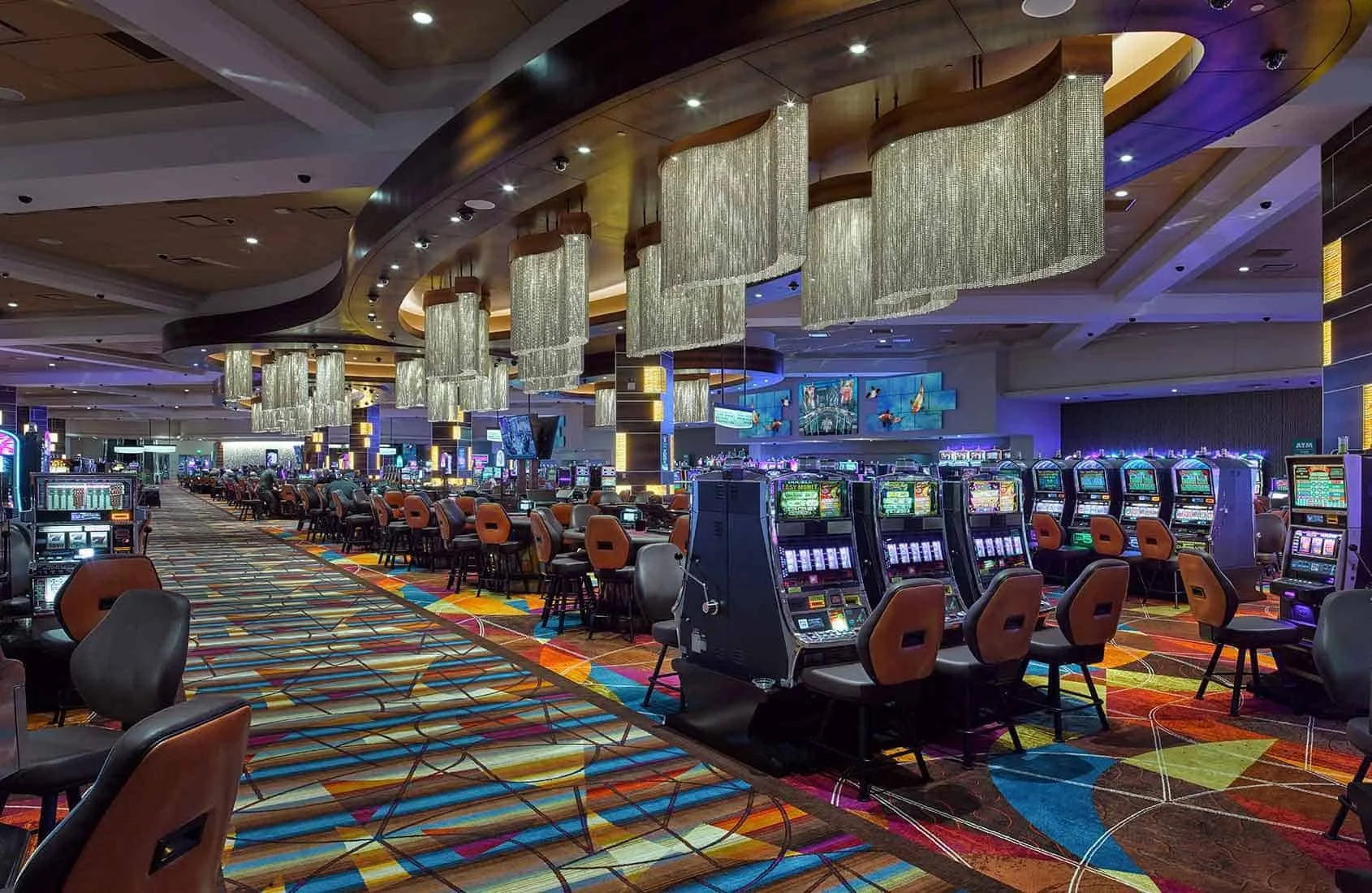 A large casino gaming floor with rows of slot machines, colorful carpet, and elegant hanging chandeliers in a modern setting.
