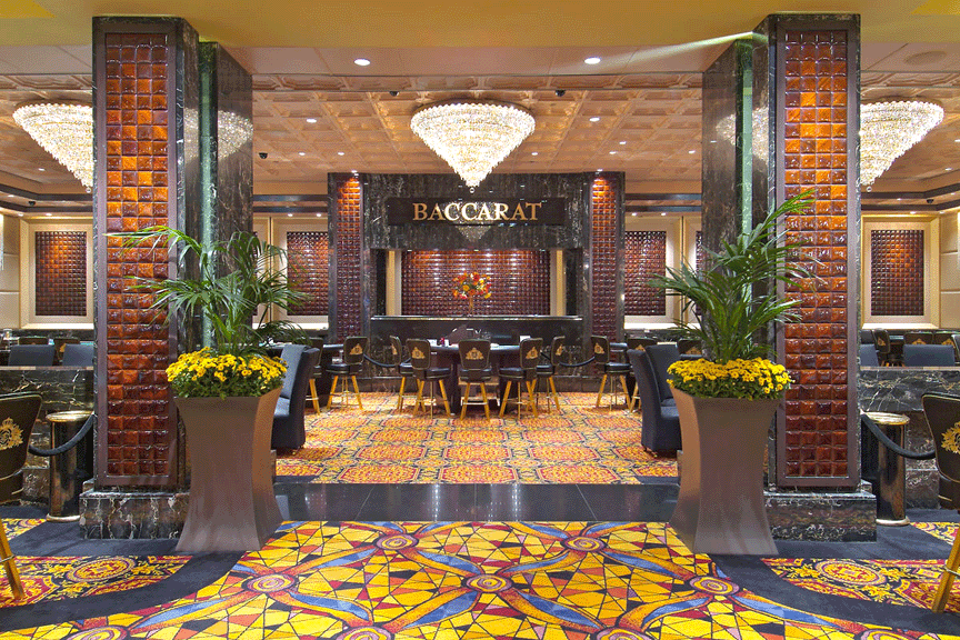 Luxurious casino interior with baccarat table at center, surrounded by chairs, ornate chandeliers overhead, decorative plants, and a vibrant patterned carpet.