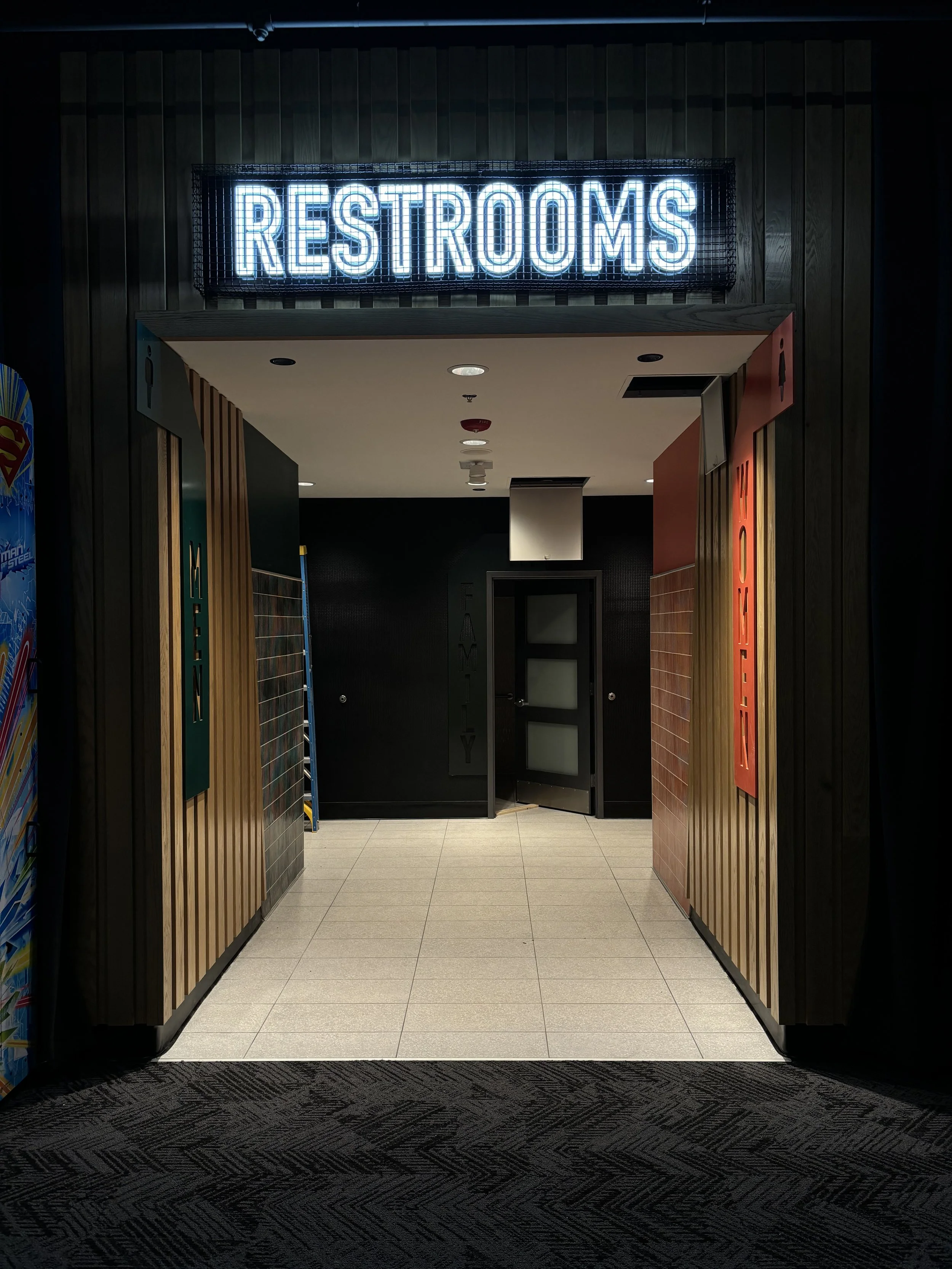 Entrance to a public restroom with a blue illuminated "Restrooms" sign overhead, dark wood paneling on the walls, and two signs indicating men's and women's restrooms on the sides.