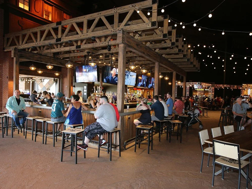 People sitting at a bar with TV screens, surrounded by string lights, in a rustic-style venue with exposed wood beams and brick walls, some socializing and others watching the screens.