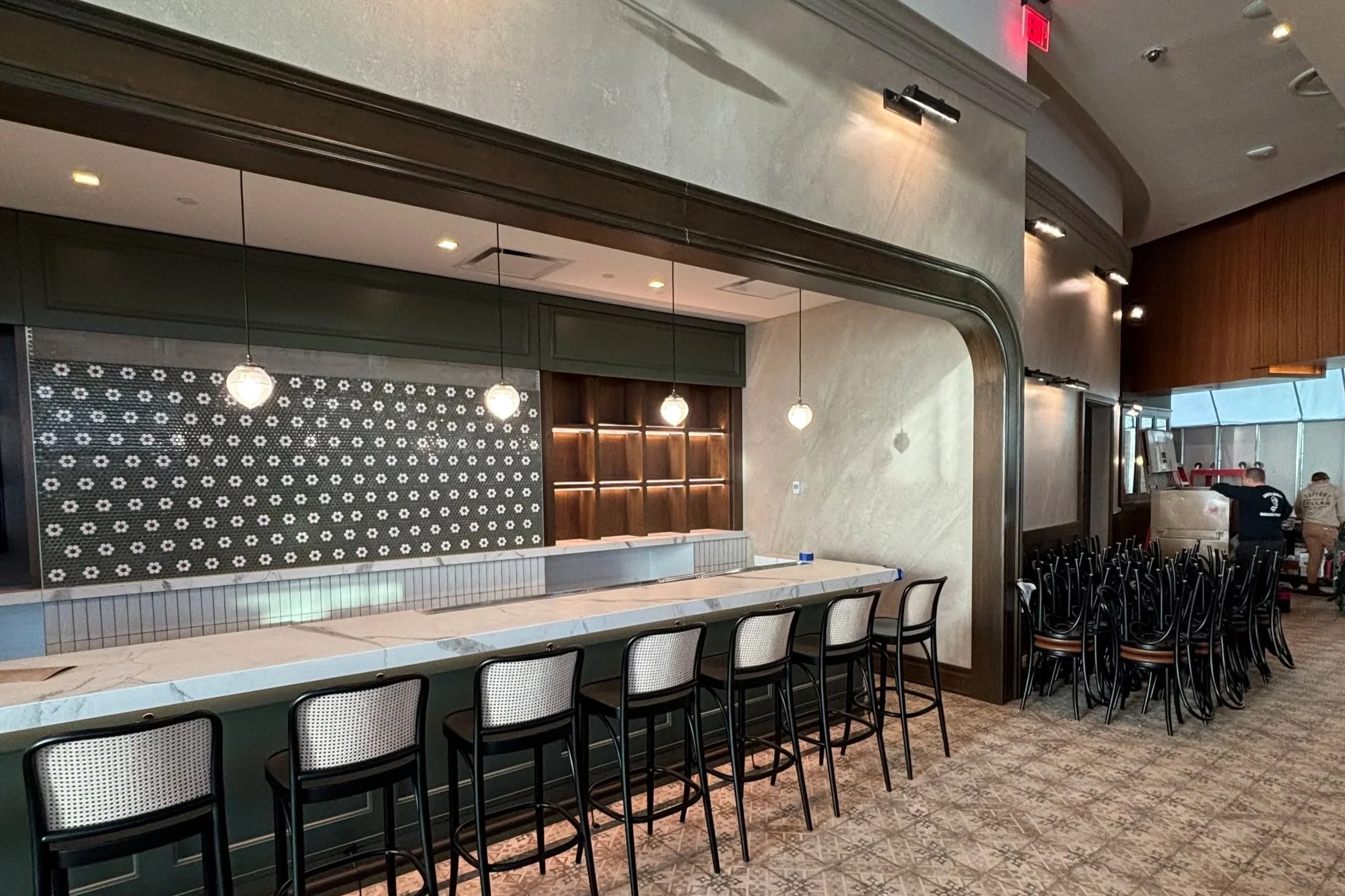 Empty bar area with high chairs and hanging pendant lights in a modern interior with patterned tile flooring and a decorative wall.