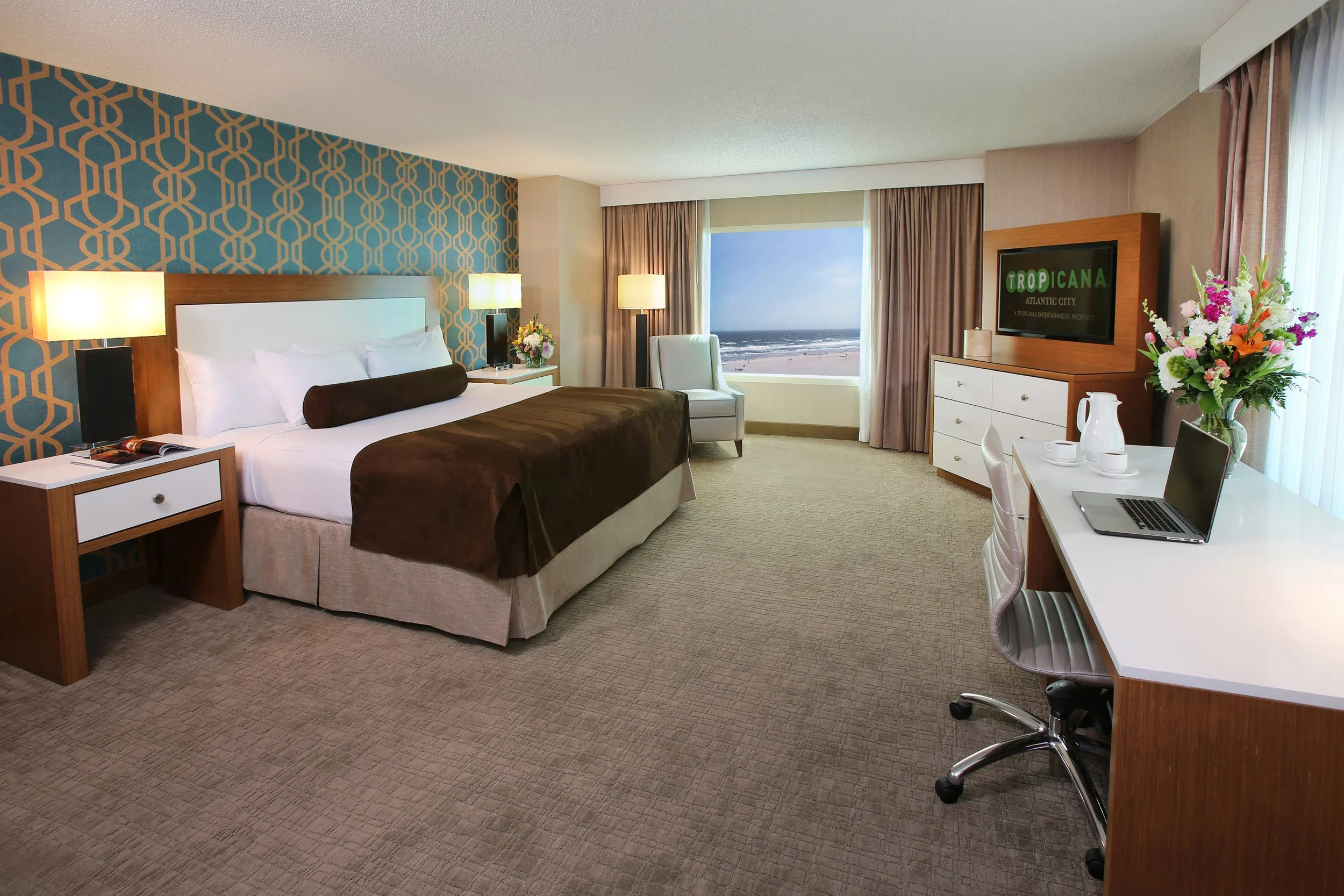 Hotel room with a queen-sized bed, bedside tables with lamps, a white armchair, a dresser with a TV, a desk with a laptop and flowers, and a large window overlooking the beach and ocean.