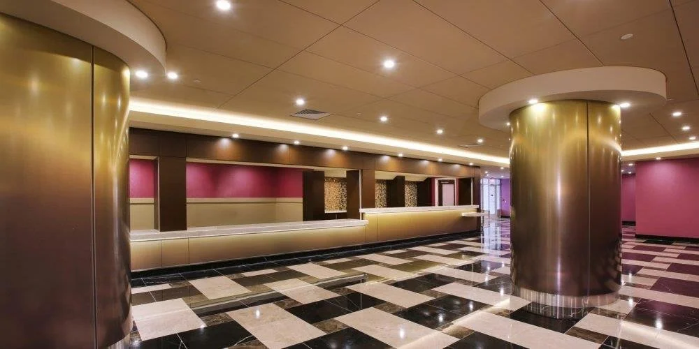 Empty hotel lobby with check-in counter, pink and beige walls, black and white checkered tile floor, large cylindrical gold-colored columns, and ceiling recessed lighting.