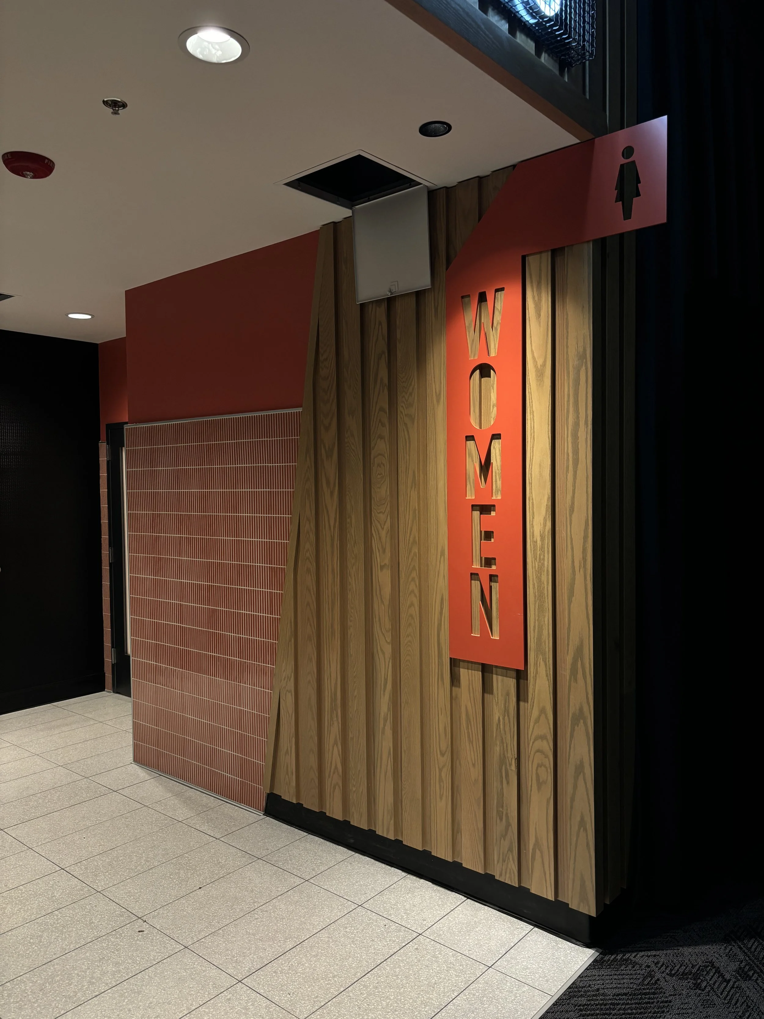 A wall with a vertical red sign that says 'WOMEN' and an arrow pointing right, indicating the location of the women's restroom.