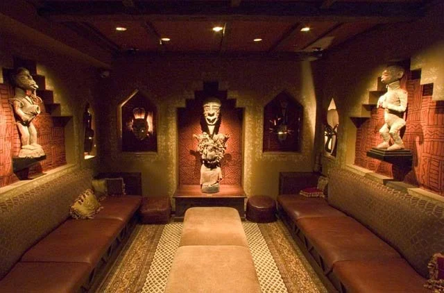 A dimly lit room with Moroccan or Middle Eastern decor, including carved wooden alcoves and sculptures of children on the walls. There are long cushioned couches along the sides and a central ottoman. An ornate vase with flowers is displayed in a nic