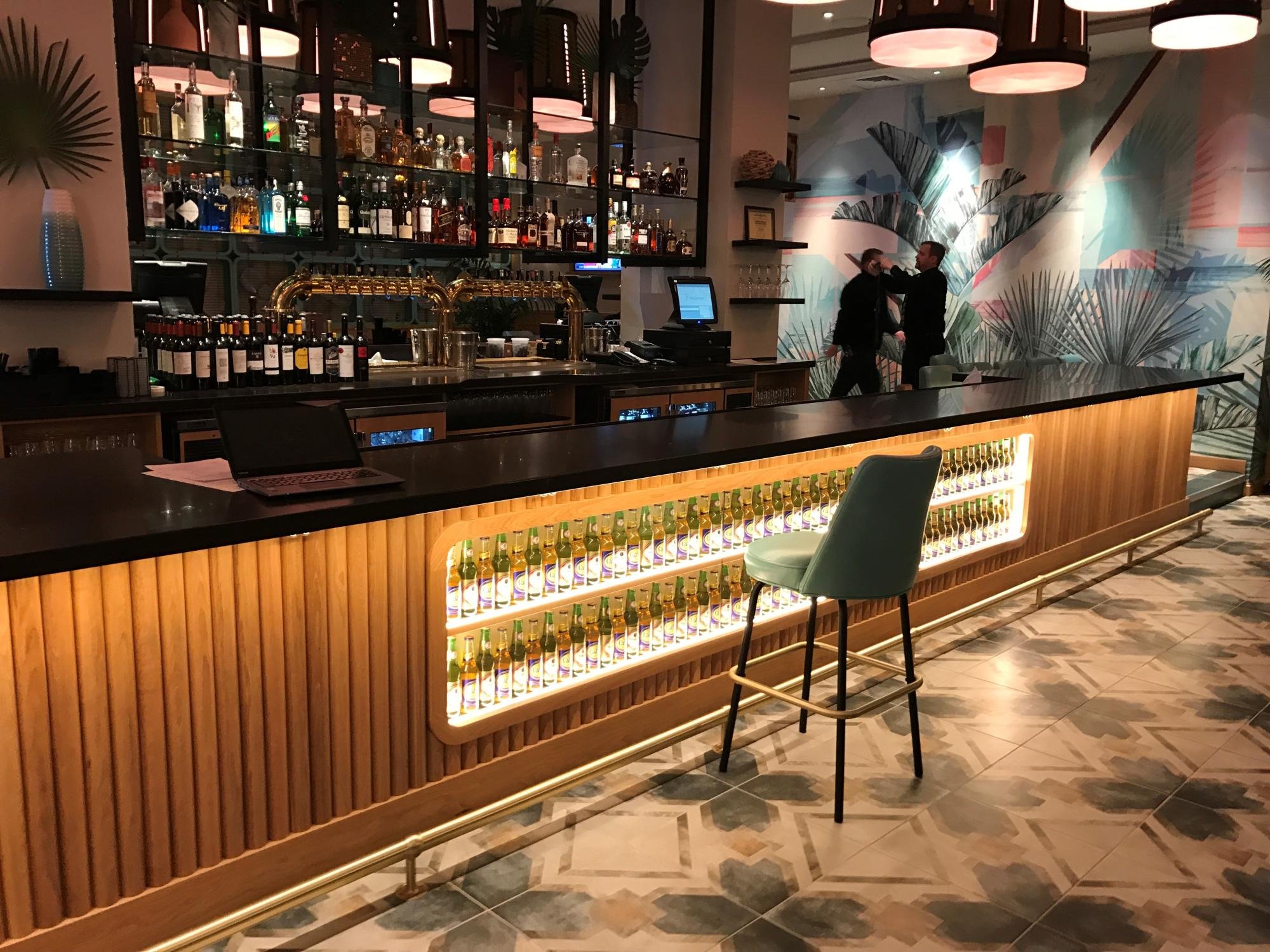 Interior of a modern bar with a black counter, a backlit display of bottled beverages, and a decorative tropical mural wall in the background. Two people are talking on the far side of the bar.