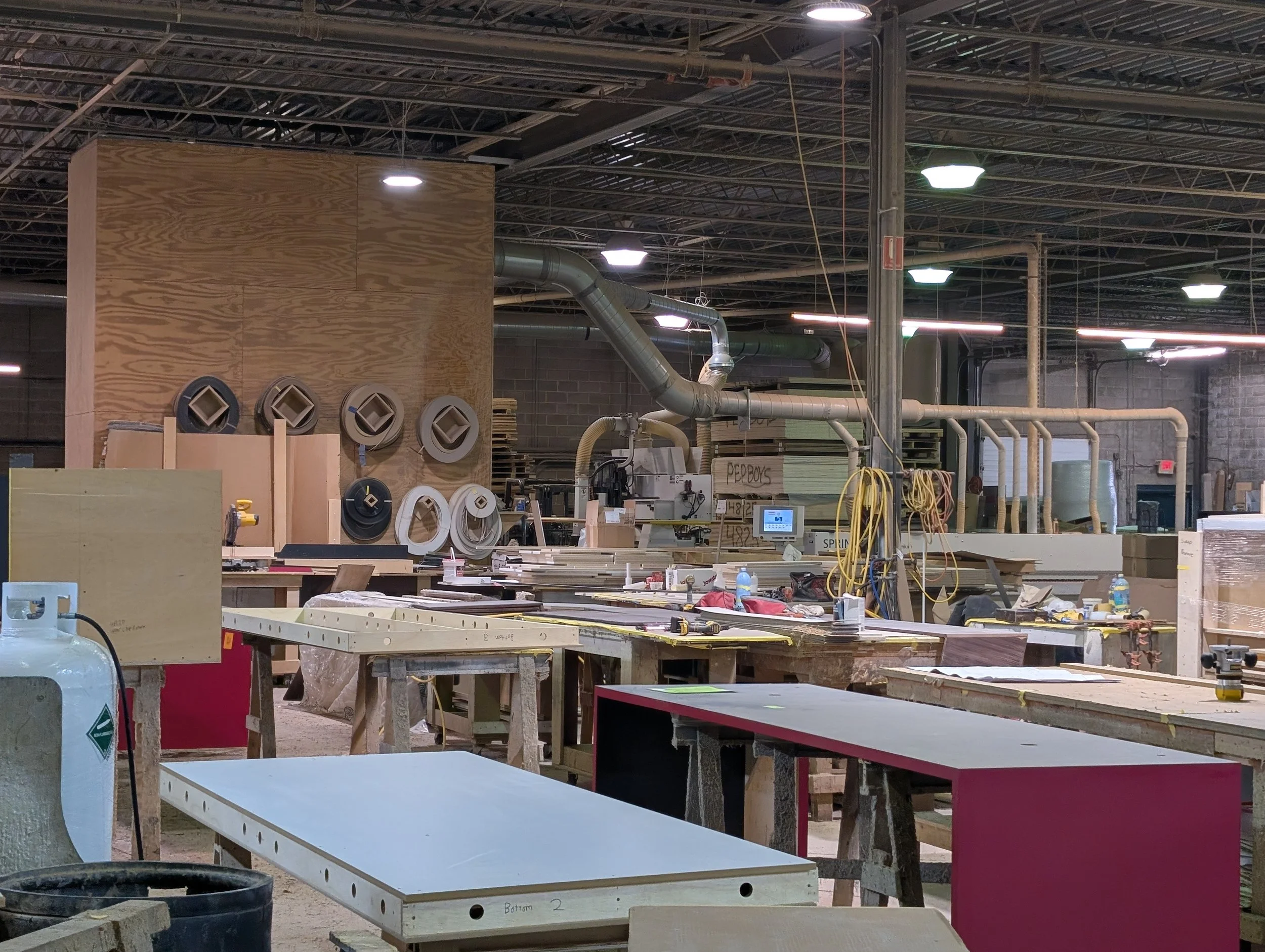 Woodworking shop with tables, tools, and wood pieces, industrial ventilation system overhead, and sewing/finishing area in background.