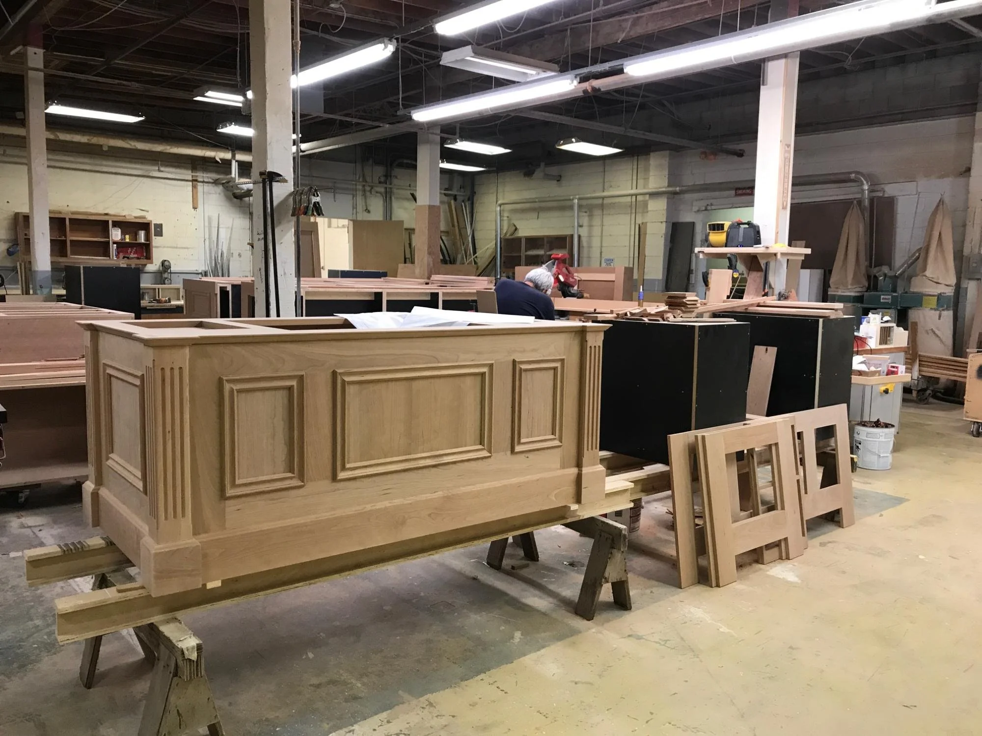 Inside a woodworking workshop with a large, unfinished wooden cabinet and several other wooden pieces being crafted.