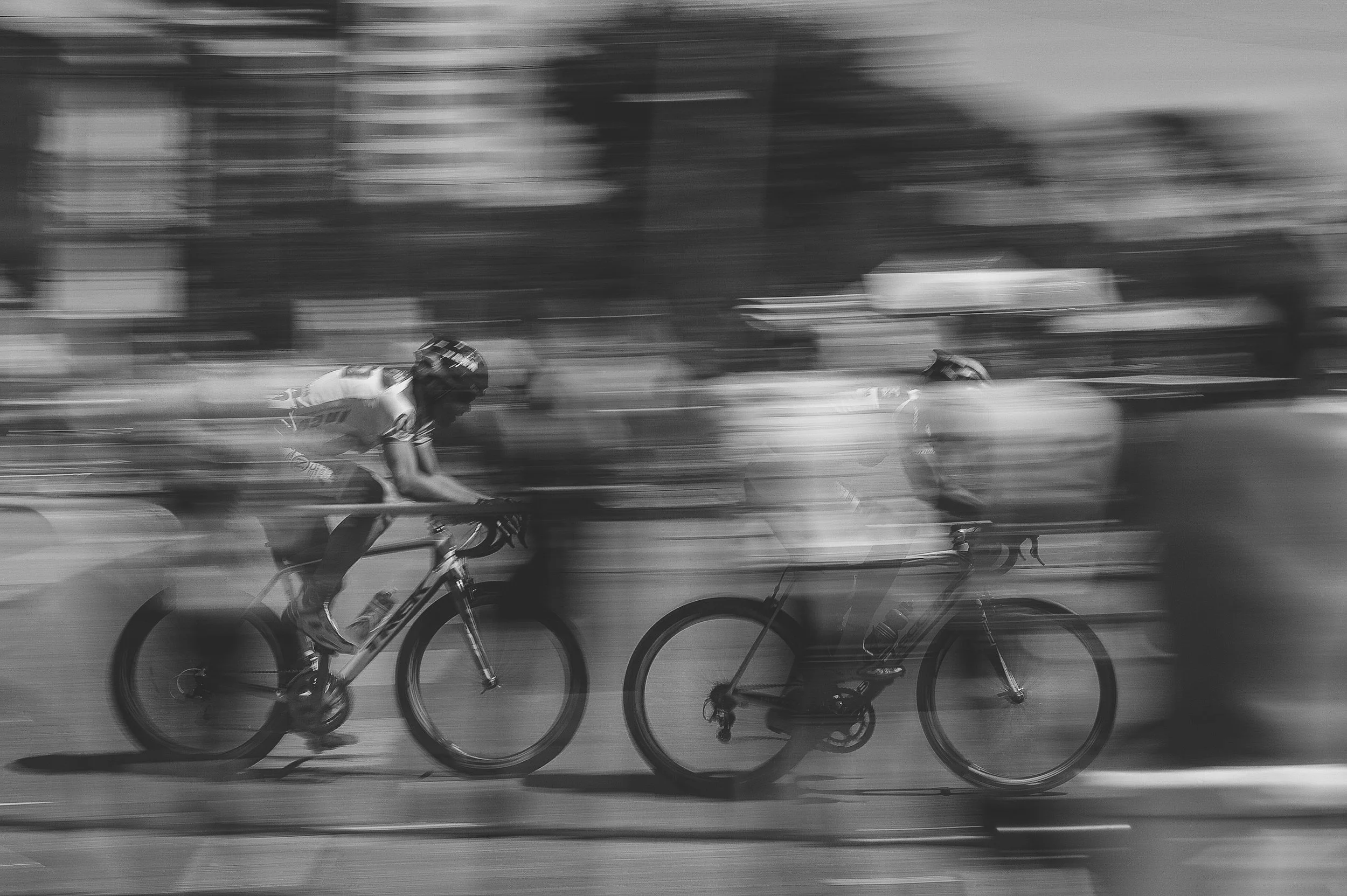 Black and white photo of two cyclists racing, with motion blur background and wheels, wearing helmets and sportswear.