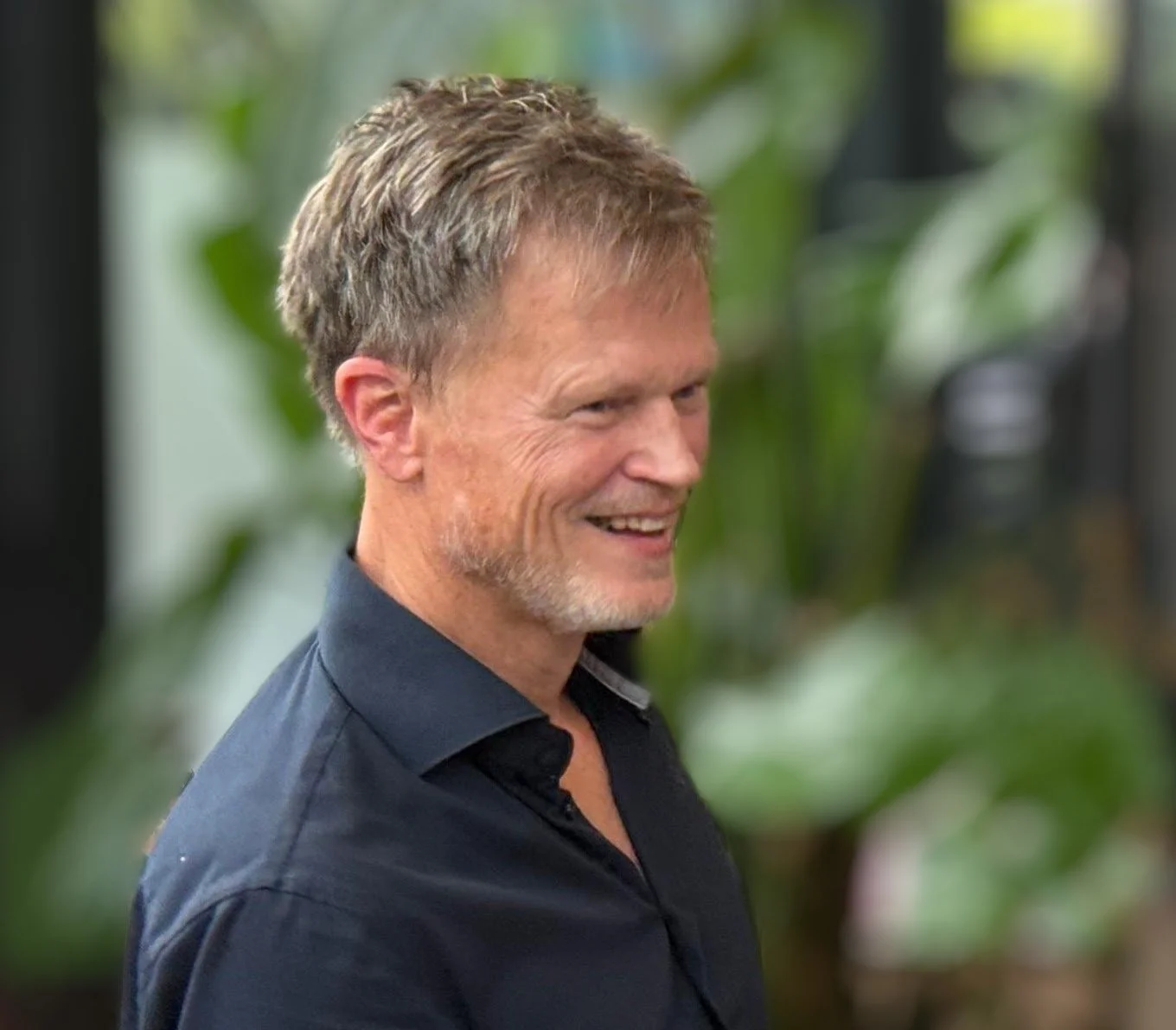 A middle-aged man with short, light brown hair and a beard, smiling, wearing a dark collared shirt, standing indoors with green plants in the background.