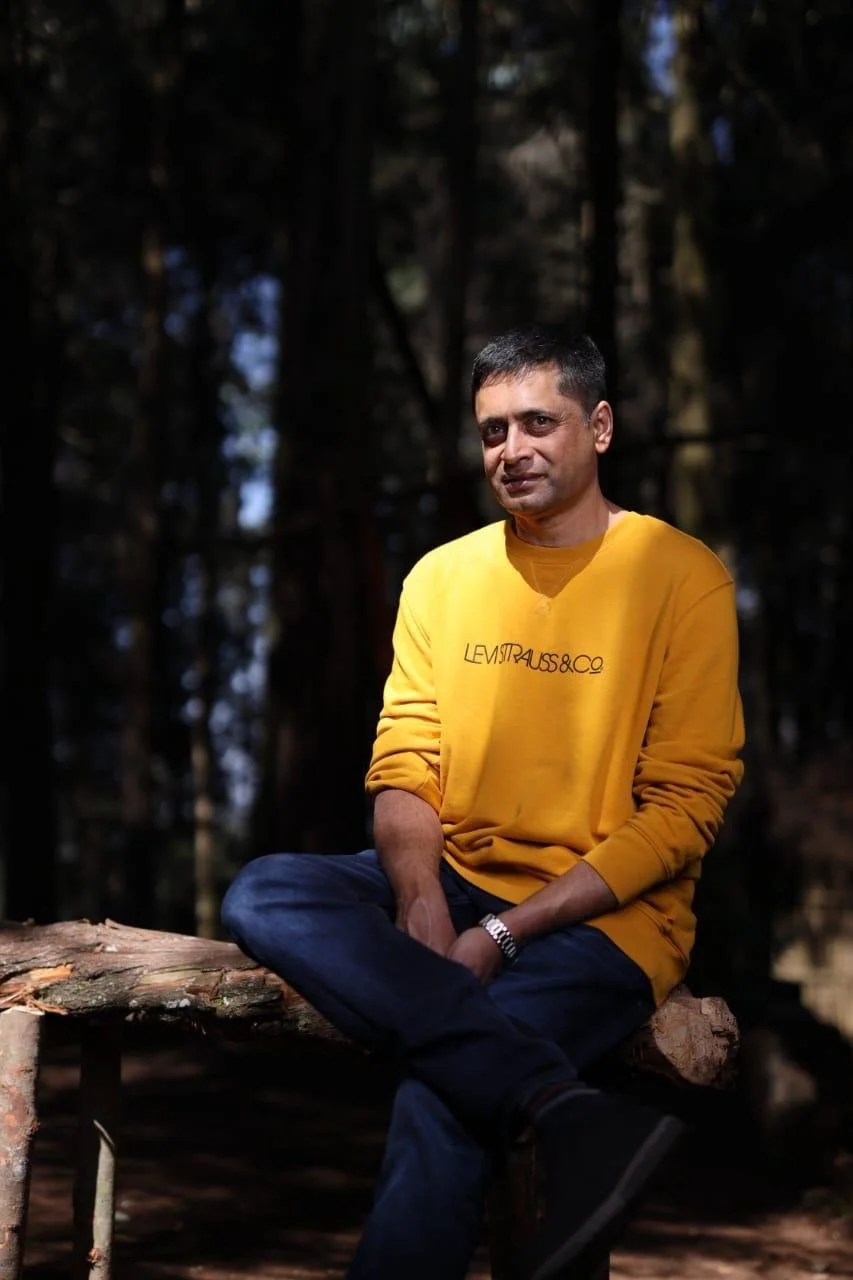 A man in a yellow sweatshirt with "Levi Strauss & Co" logo, sitting on a log in a forest with dark trees in the background.