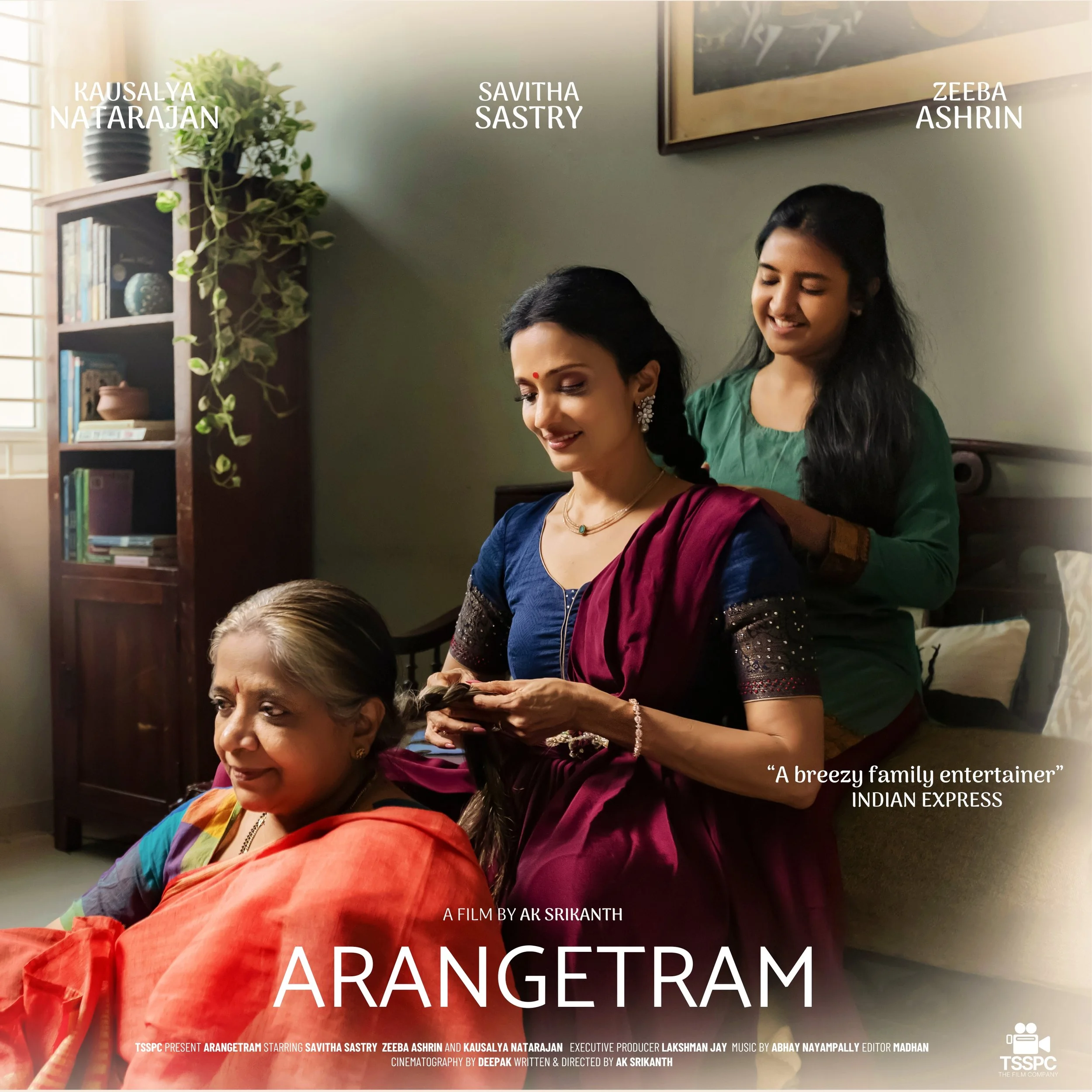 Three women in a cozy room, with one woman sitting on a bed and two women standing behind her, styling her hair. The woman sitting is wearing a colorful traditional saree, and the two standing women are also dressed in traditional attire. A bookshelf with books and decorative items is seen in the background, along with a framed picture on the wall. The scene appears warm and familial, with sunlight streaming in from a window.