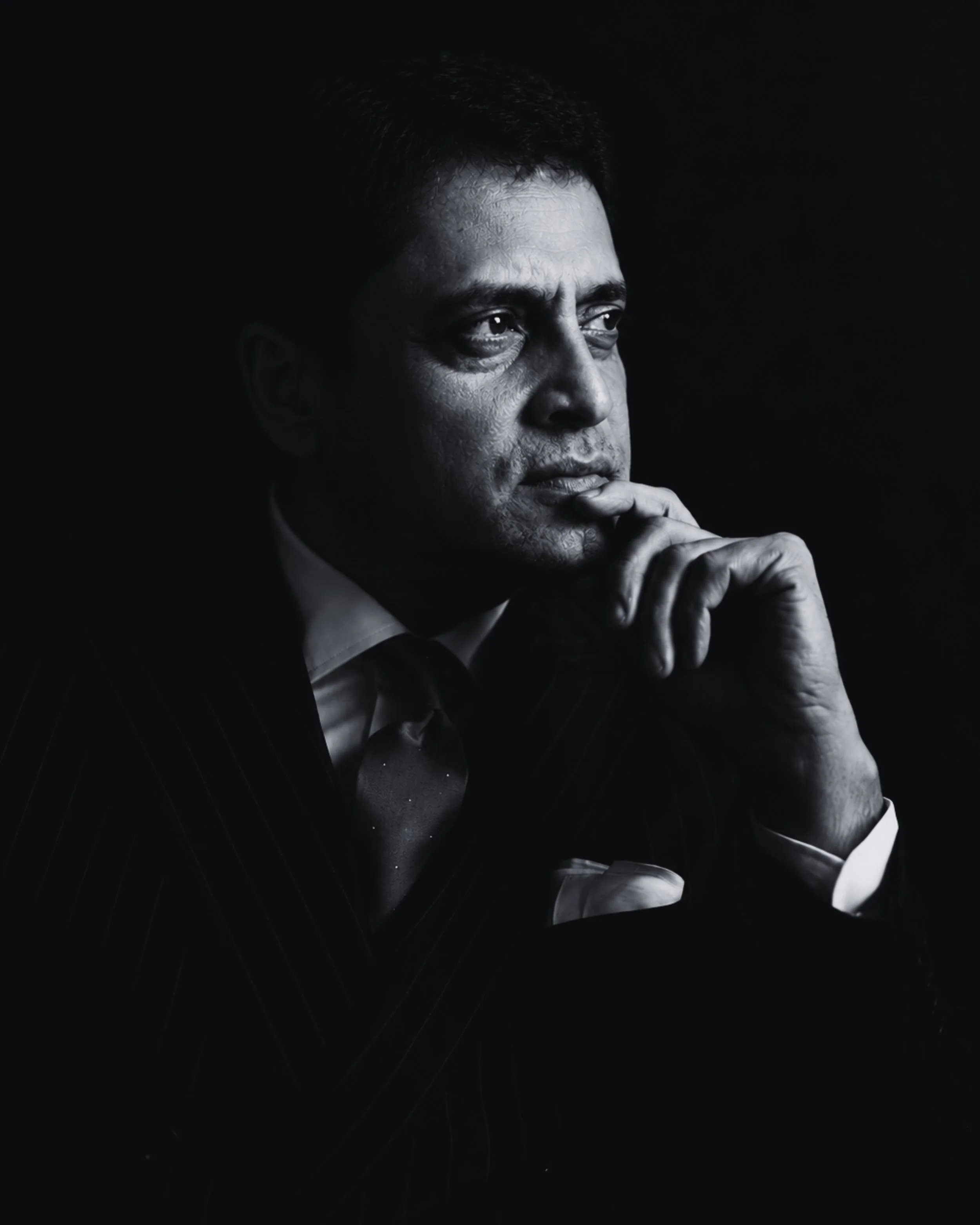 A black and white portrait of a man with contemplative expression, wearing a suit and tie, resting his chin on his hand, against a dark background.