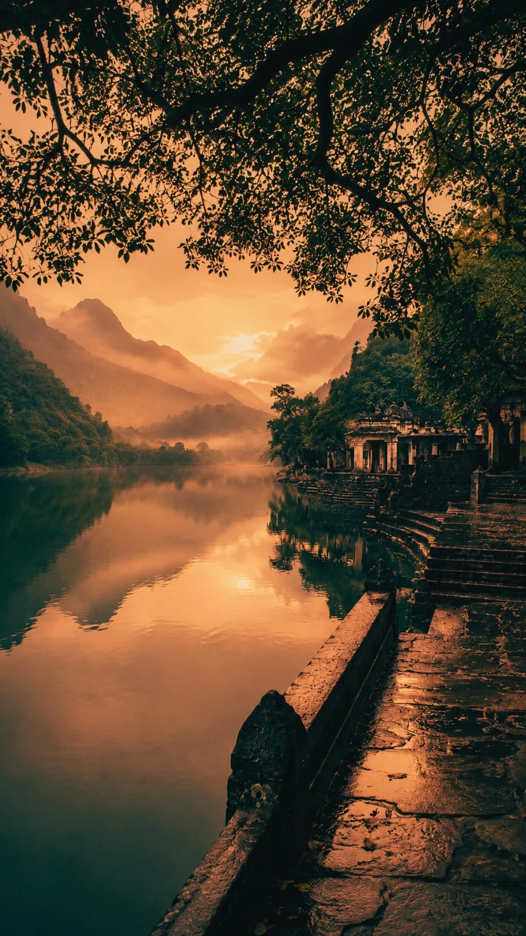 Sunset over a river with mountains in the background, trees framing the scene, and ancient stone steps and buildings along the water's edge.
