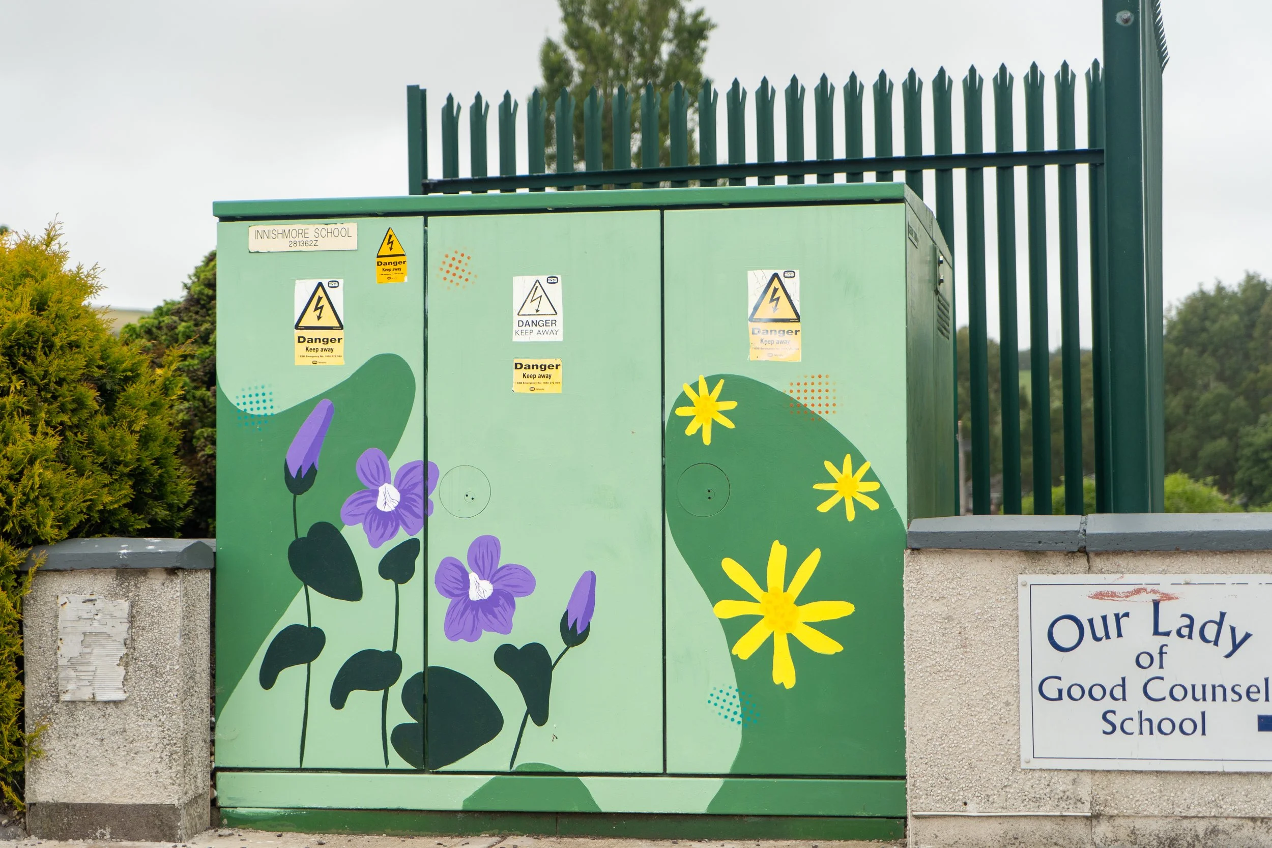 Front image of utility box painted with flowers. A fence is behind the box and a school sign on the adjacent wall.
