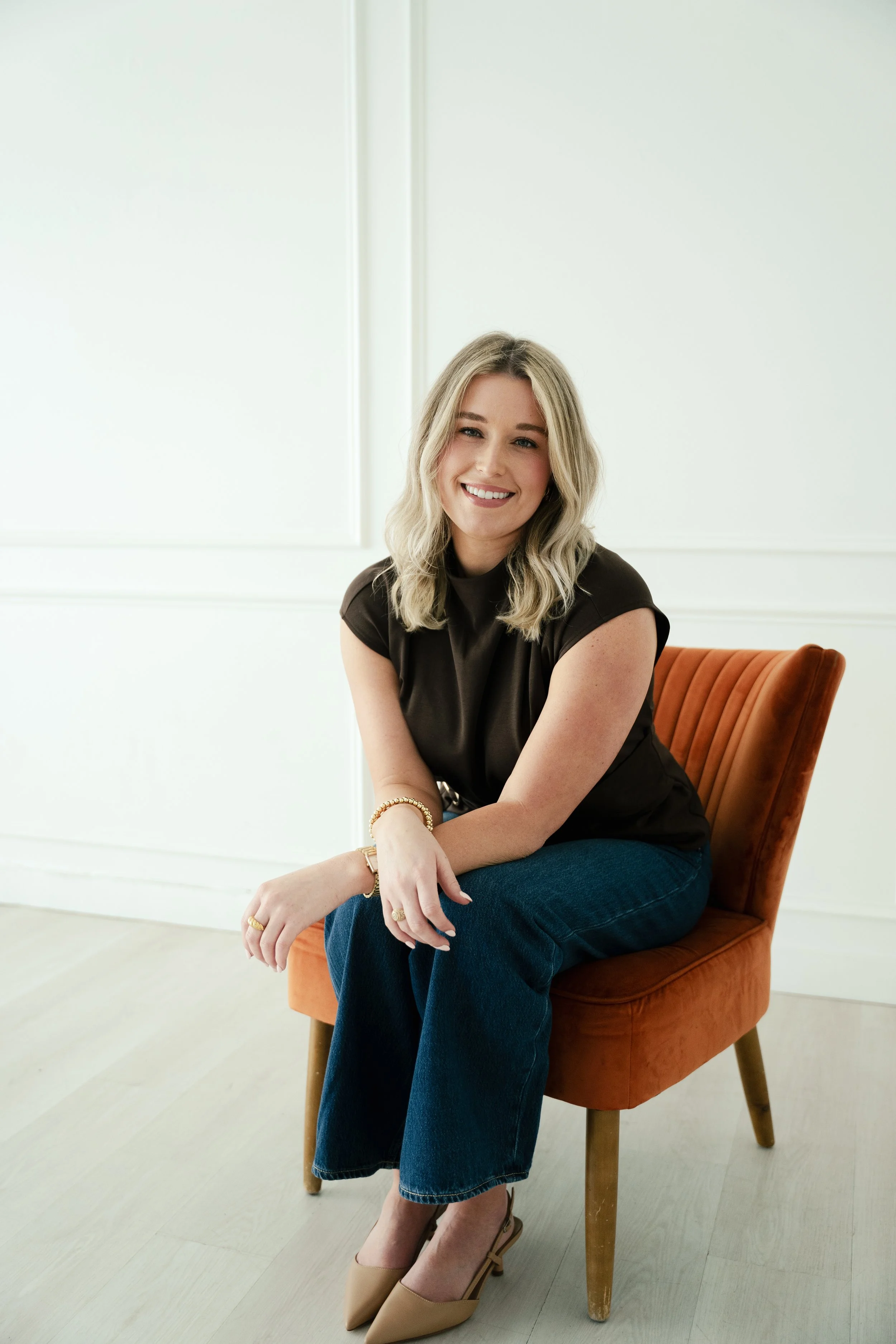 A young woman with blonde hair sitting on an orange velvet chair in a bright, minimal room. She is smiling and wearing a black top, jeans, beige high heels, and jewelry.