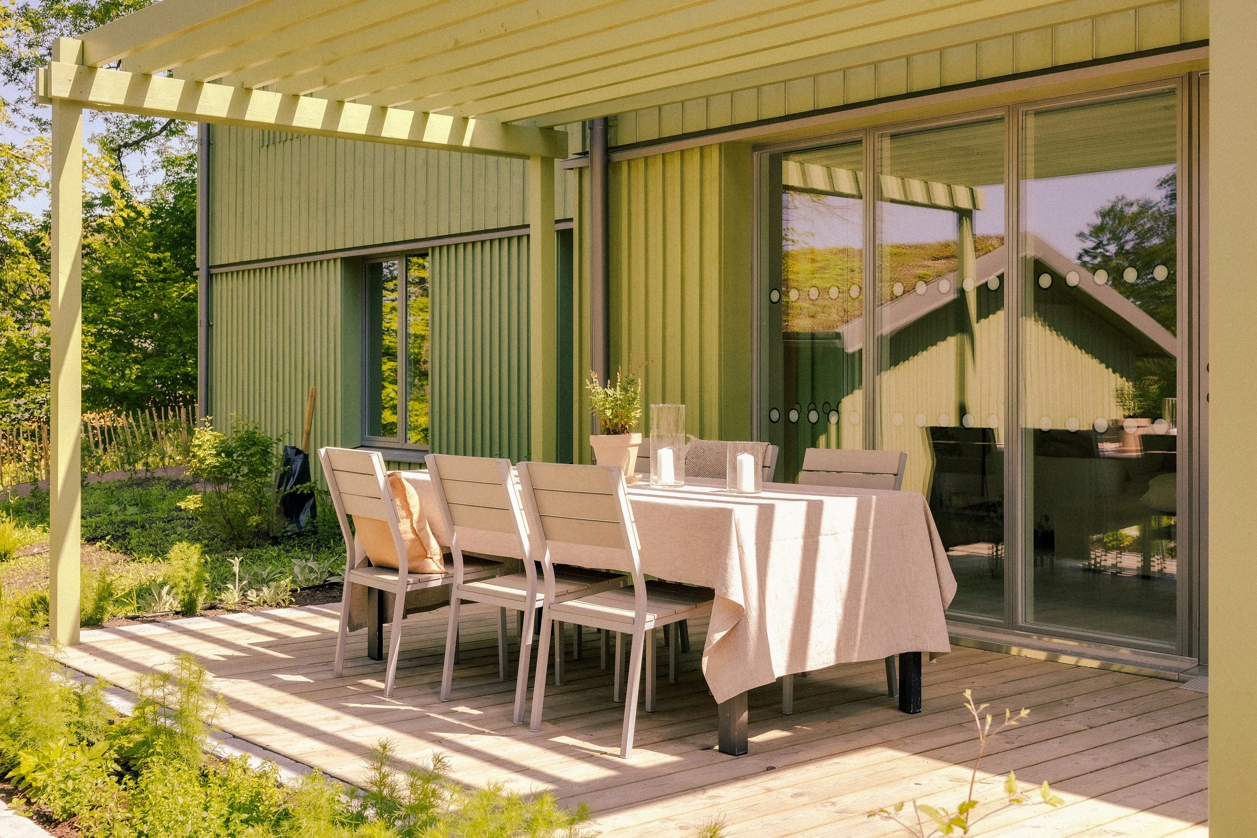 Utomhus matbord med stolar på trädäck utanför ett grönt hus med stora glasfönster och en pergola, omgiven av växter och träd.