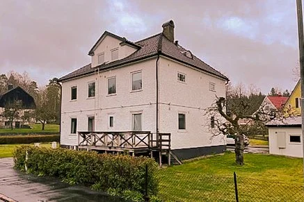 En vit tvåvåningshus med en liten träaltan och en beskuren trädgård, omgivet av ett staket.