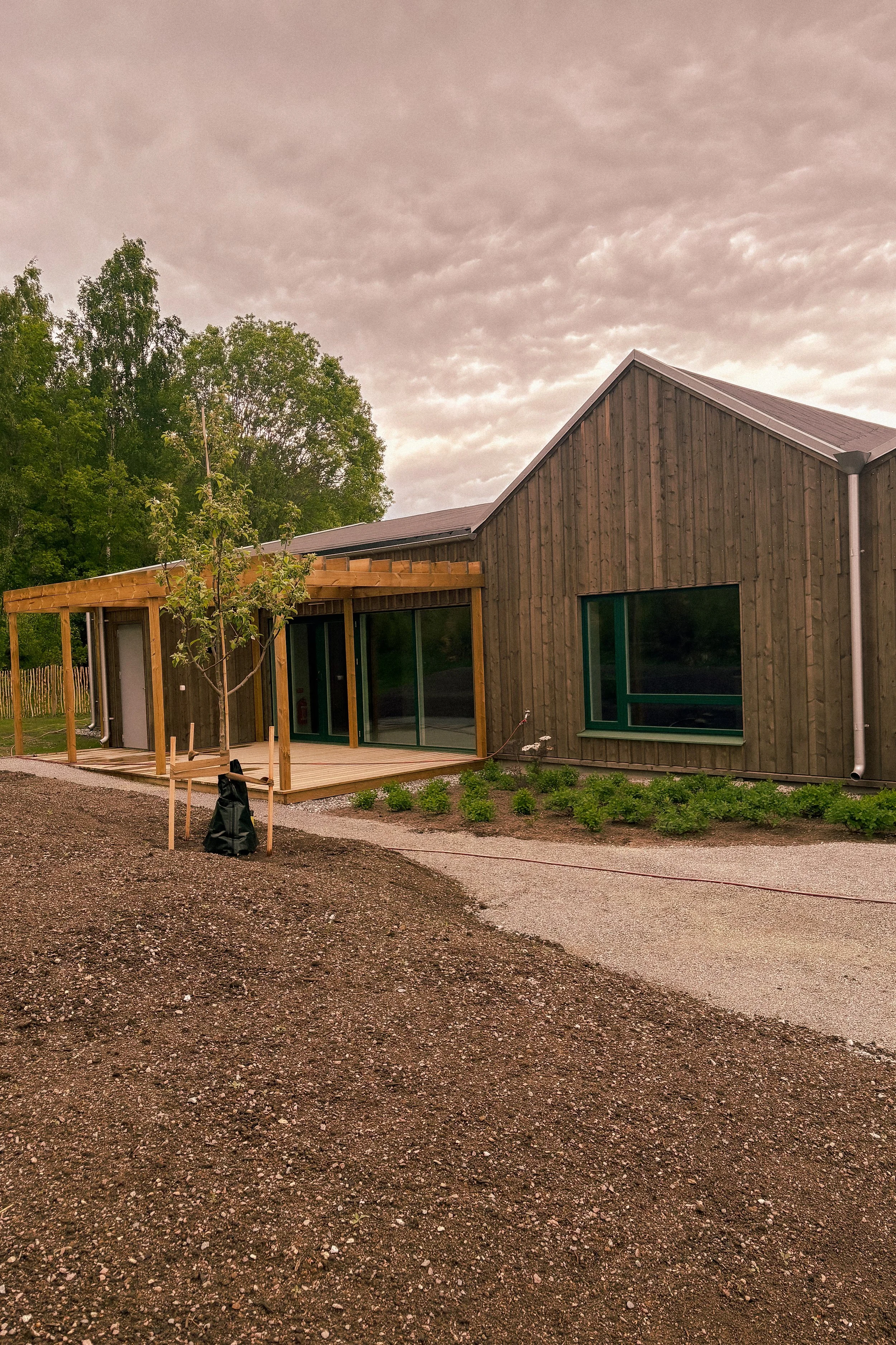 Ett nybyggt hus med träfasad och stora fönster, omgiven av gröna träd och en grusgård under en molnig himmel.