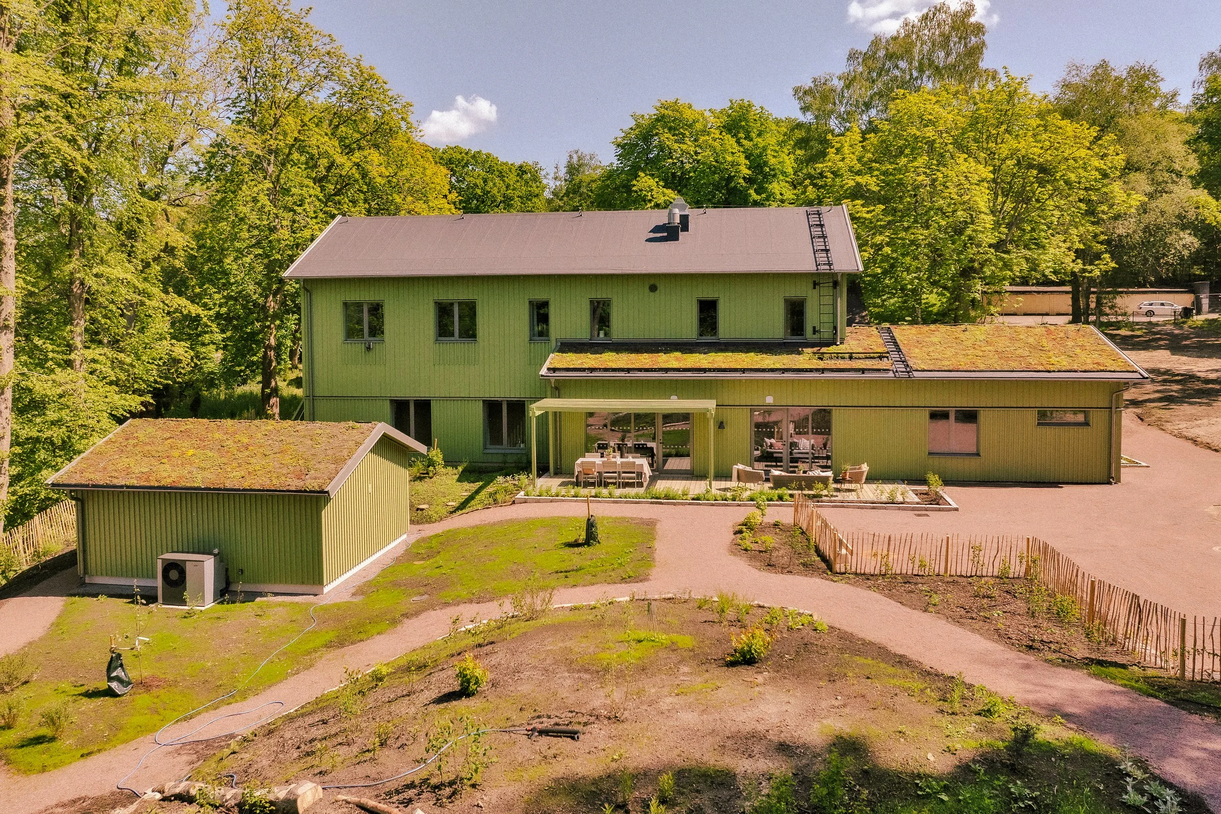 Flerbostadshus i grönt med tak som är täckt av mossa, omgiven av många gröna träd och en gräsyta framför.