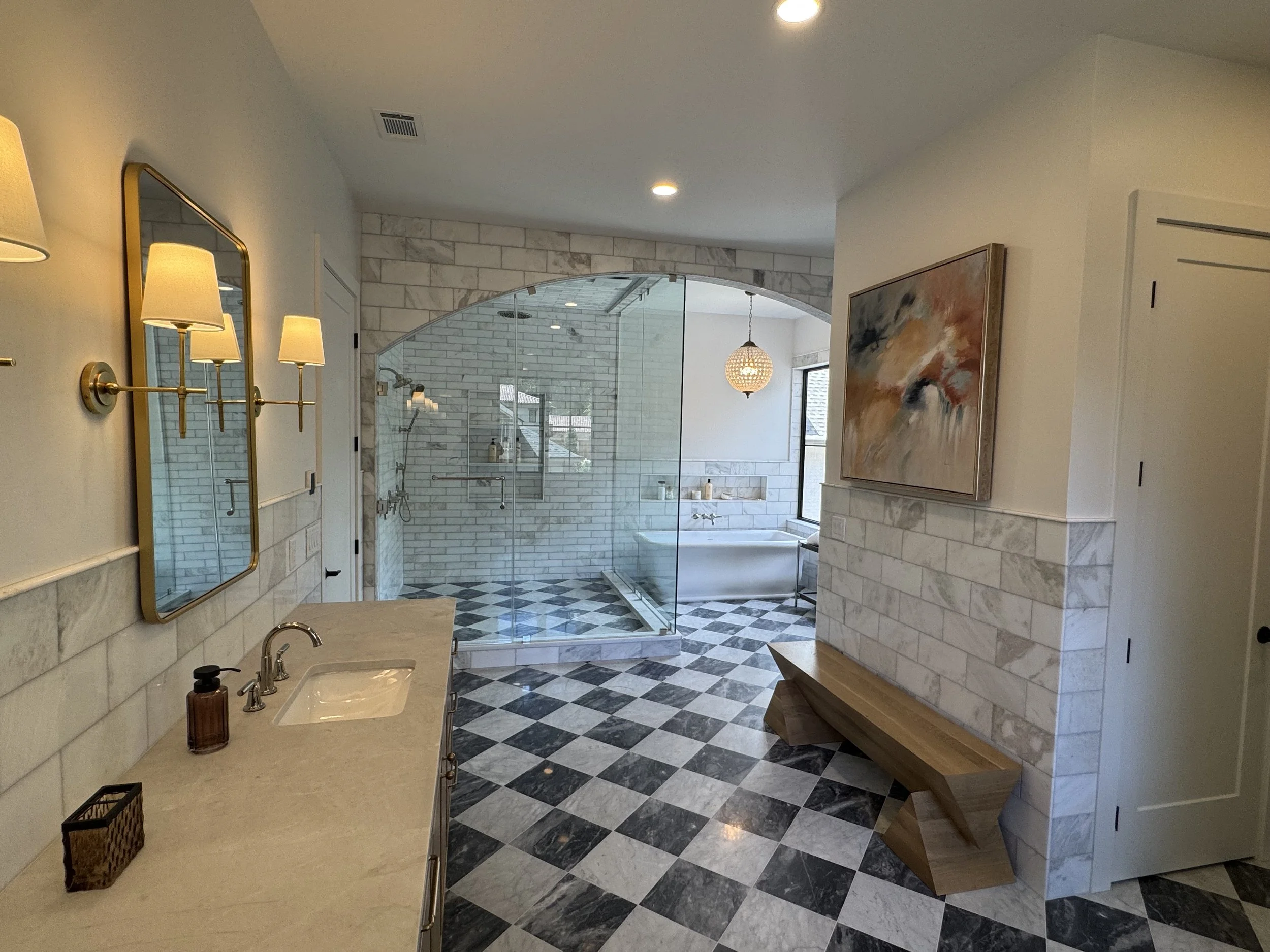 Modern bathroom with black and white checkered floor, marble walls, glass-enclosed walk-in shower, bathtub, abstract painting, wooden bench, white sink with gold fixtures, and gold-framed mirror with wall-mounted lights.