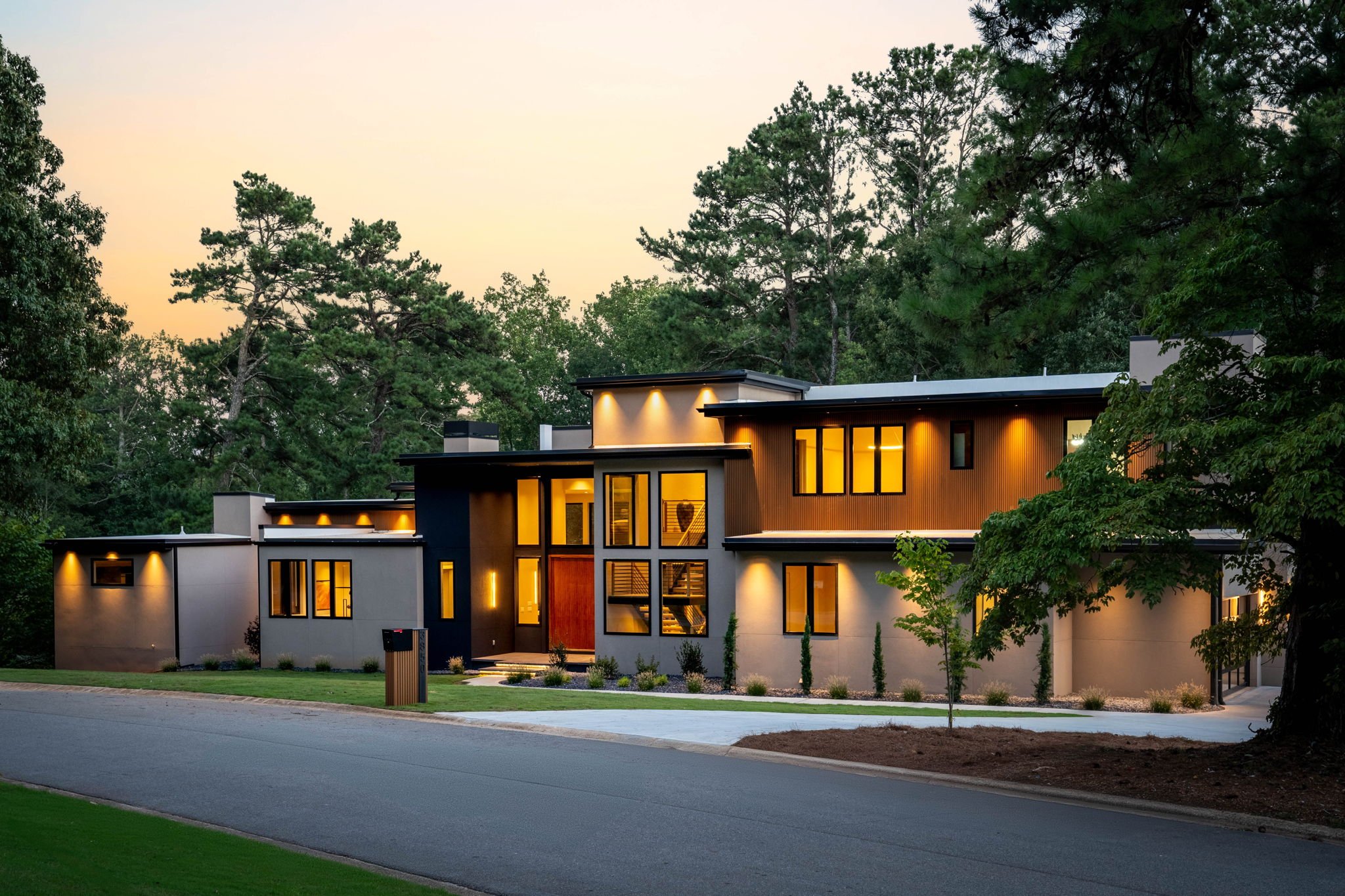 Modern house with large windows illuminated from inside, surrounded by trees and a well-maintained lawn, at dusk.