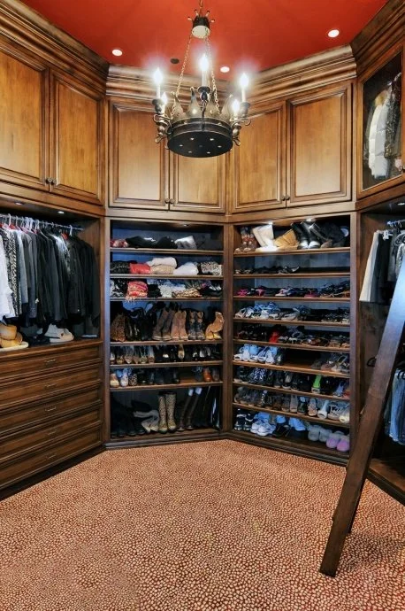 Walk-in closet with wooden shelves filled with shoes and clothes, chandelier hanging from ceiling, red ceiling, carpeted floor.