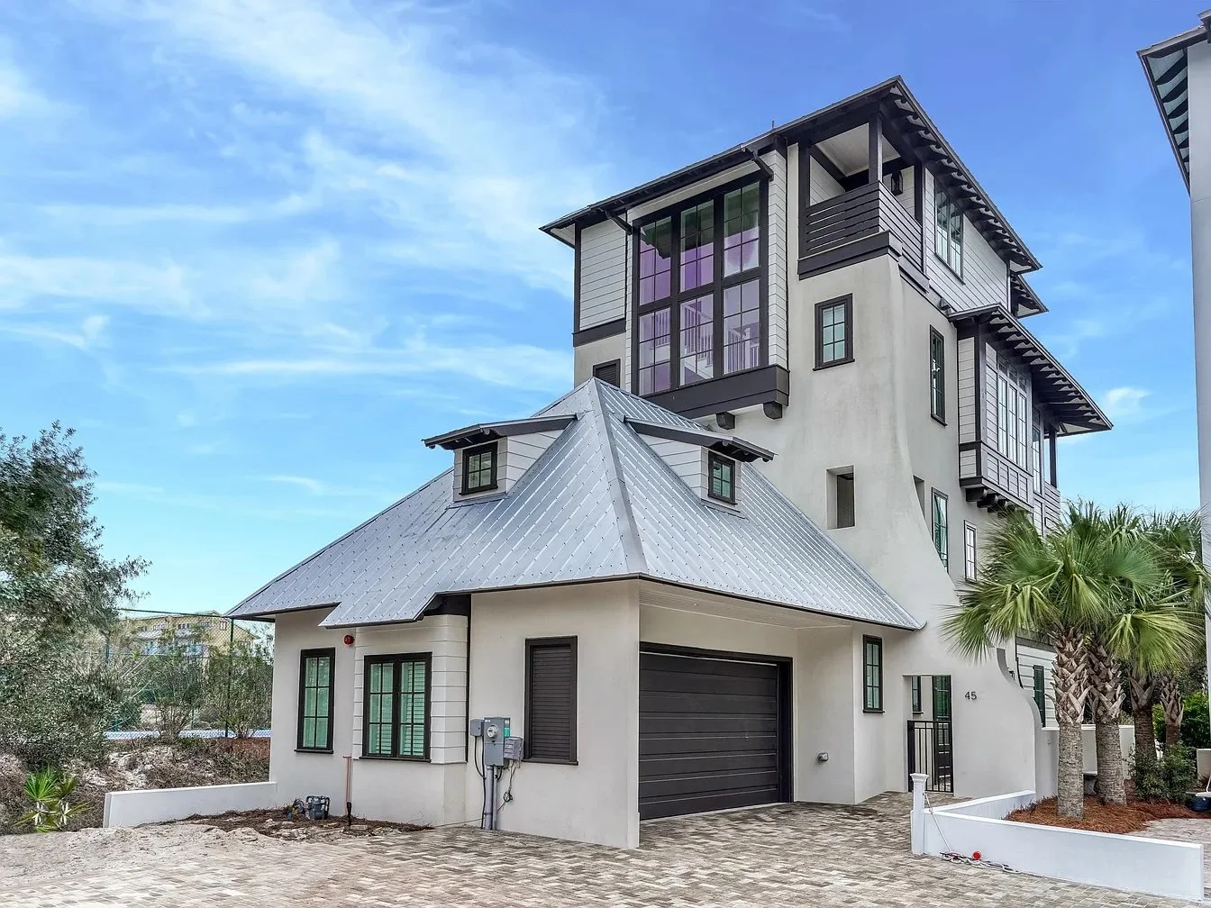 Multi-story modern house with white exterior walls, black window frames, a metal roof, and palm trees in the yard, under a partly cloudy sky.