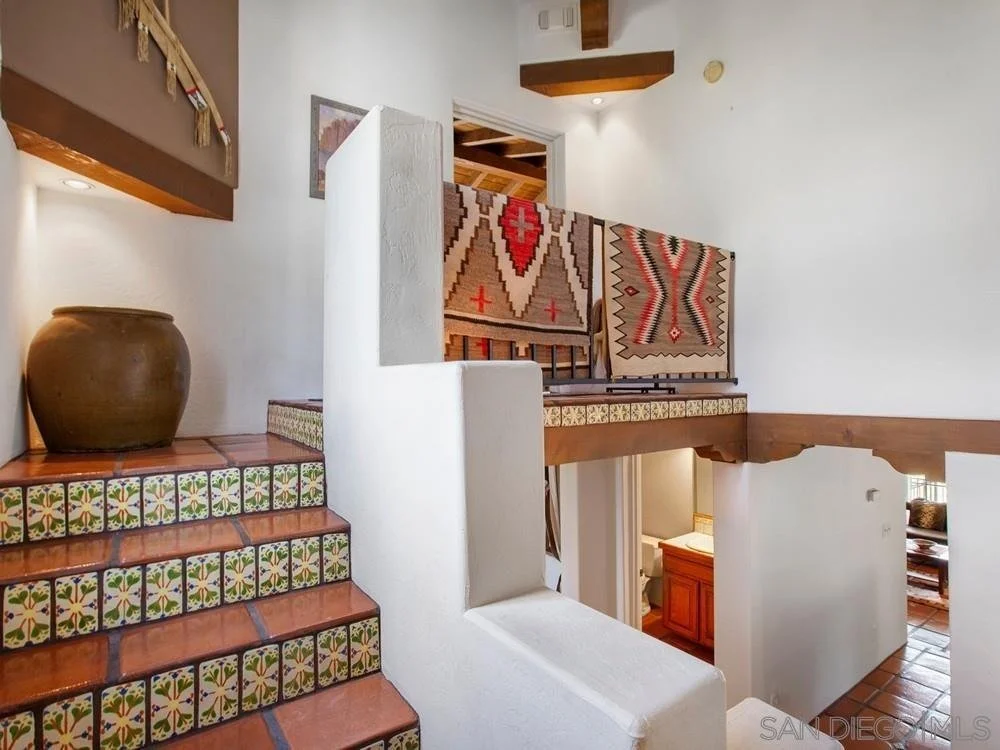 Interior view of a multi-level house with colorful southwestern style tile stairs, ceramic hand-painted tiles, a large brown pot on the landing, decorative rugs on the upper level, and wooden beams on the ceiling.