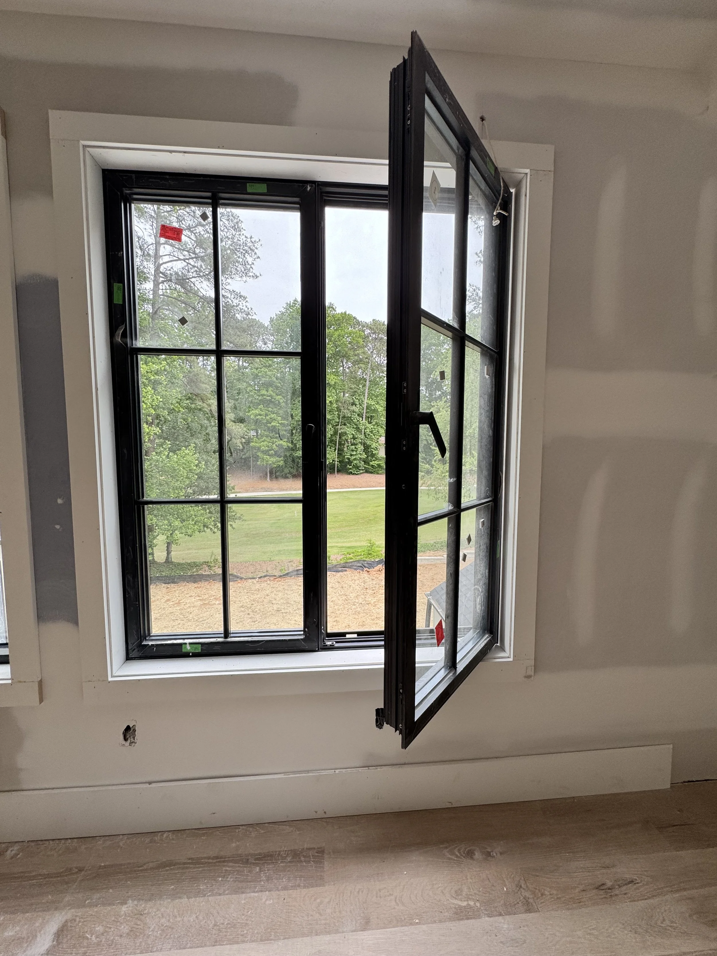 A black-framed double-hung window with multiple panes, partially open, in a residential room under construction or renovation. Outside, trees and a grassy lawn are visible.