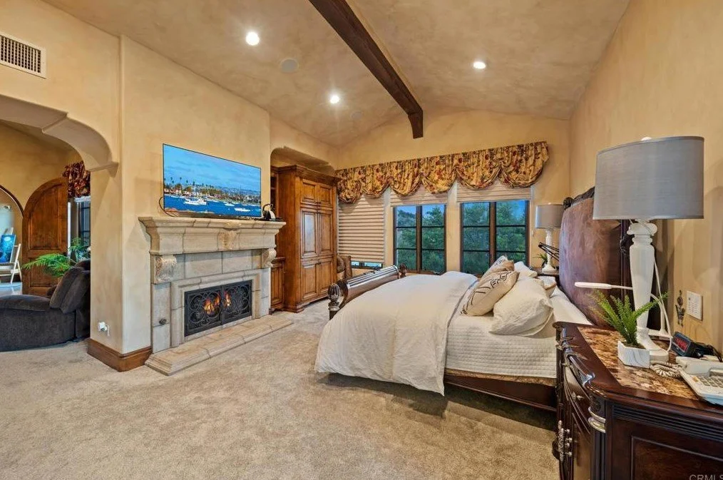A spacious bedroom with a large bed, nightstand with lamps, a stone fireplace with a TV above, large windows with floral drapes, and a wooden armoire.
