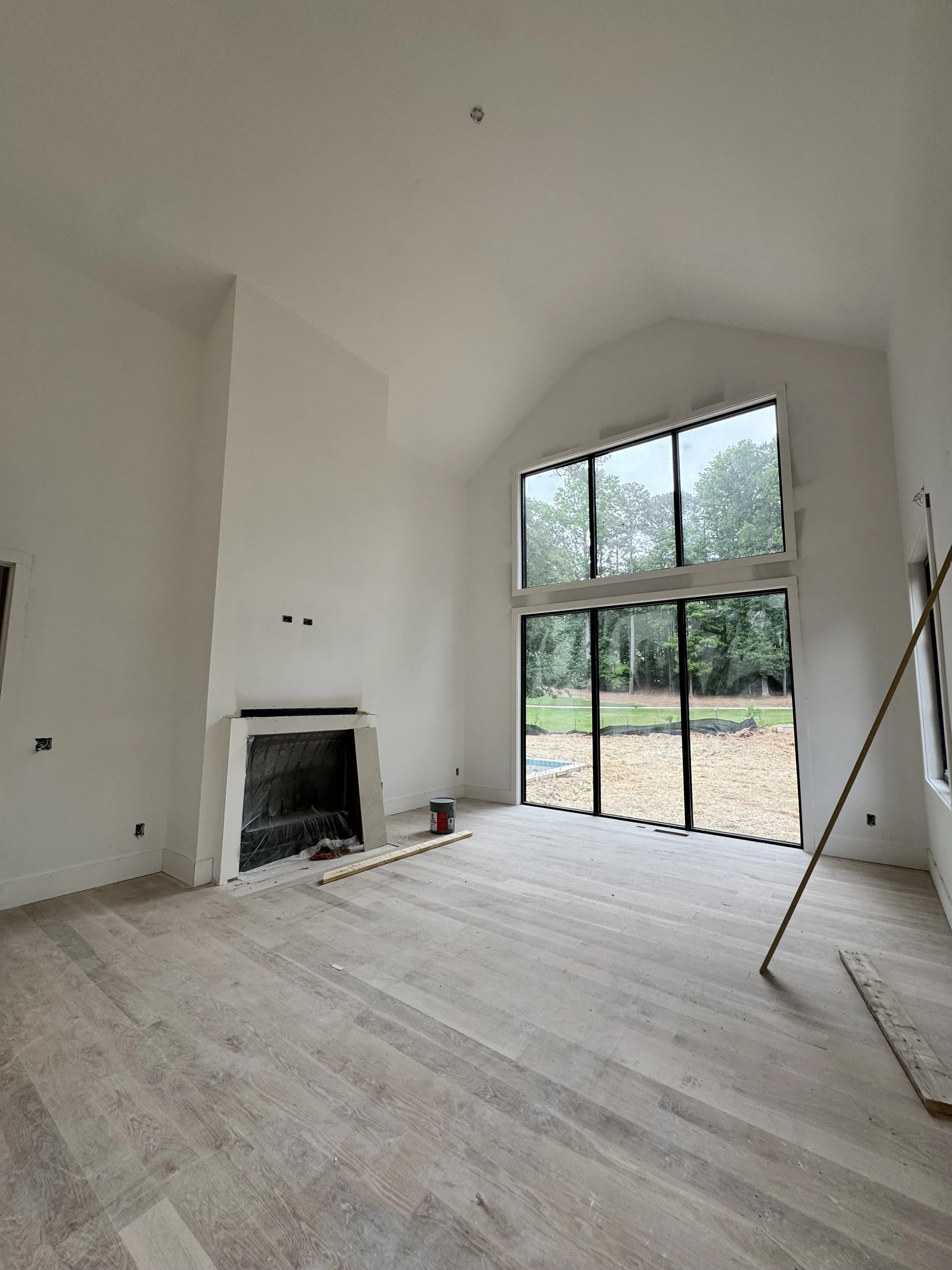 Empty living room under construction with large window showing outdoor trees and landscape, fireplace, and light wood flooring.