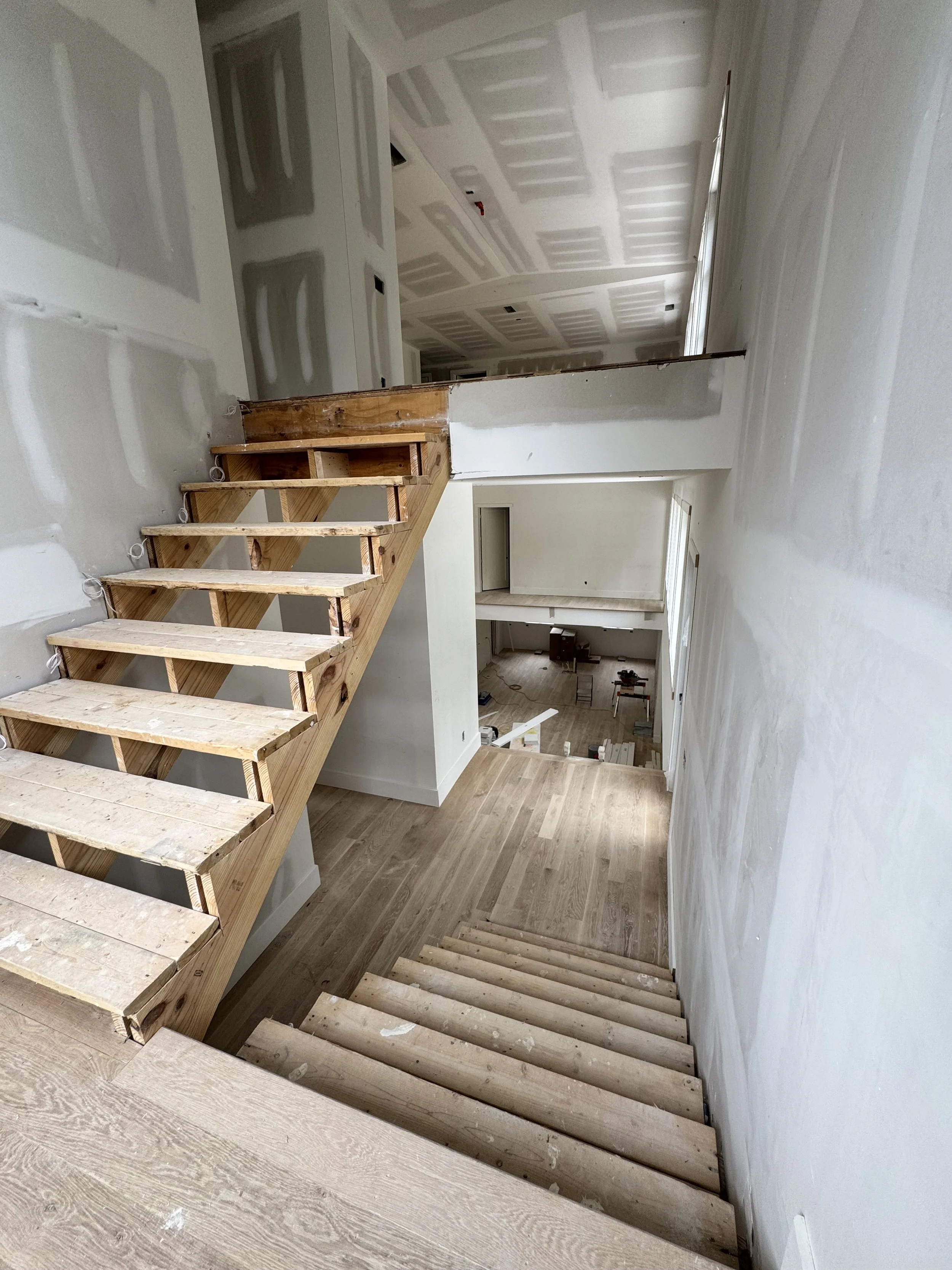 Interior view of a house under construction, showing unfinished staircase with wooden steps, drywall on the walls and ceiling, and construction tools and materials in the background.