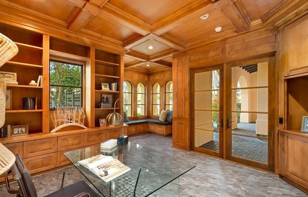 A sunlit room with wooden walls, ceiling, and built-in bookshelves, featuring a window seat with cushions, glass doors leading outside, a glass desk with magazines, and framed photos and decor on the shelves.