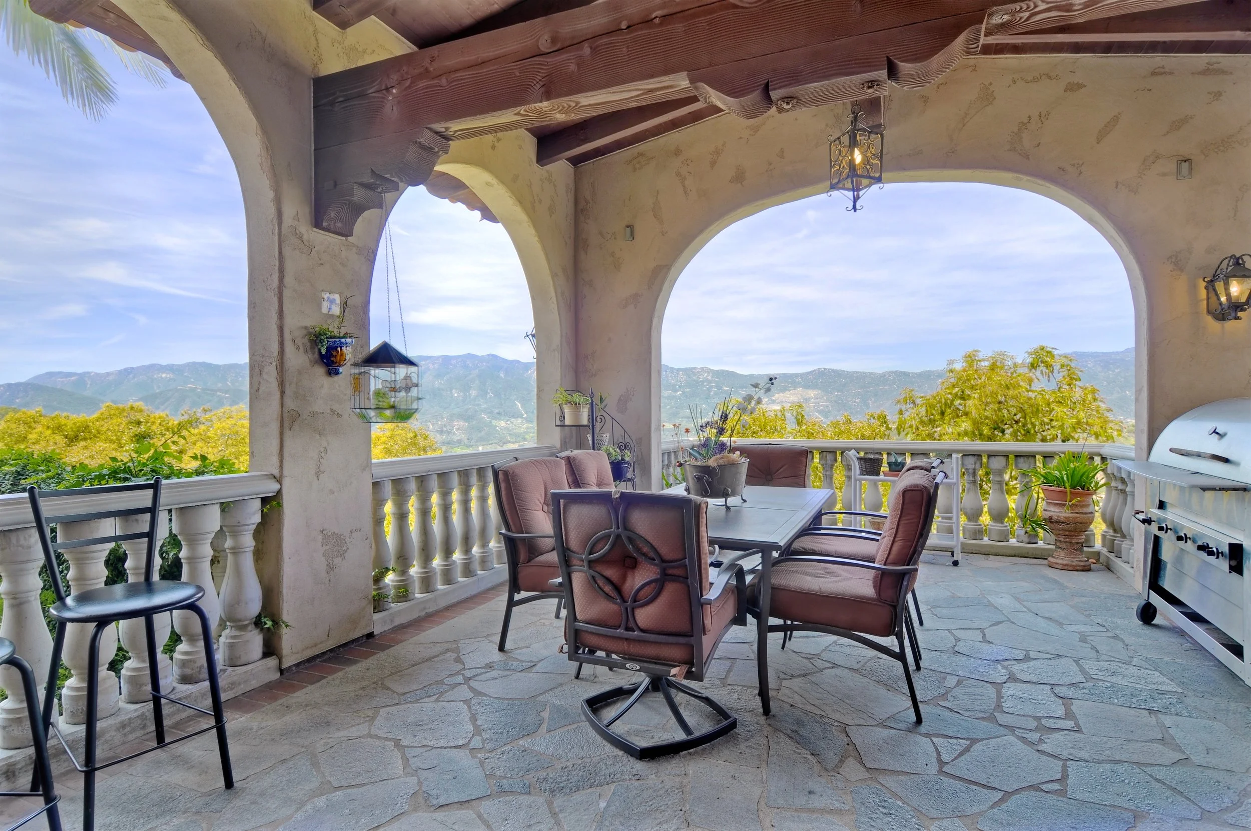 A covered outdoor patio with a dining table and chairs, with a scenic mountain view in the background, decorated with hanging plants and lantern-style wall lights.
