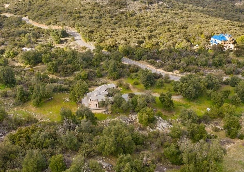 An aerial view of a rural area with trees, a winding road, a house with a blue roof, and a stone building surrounded by greenery.