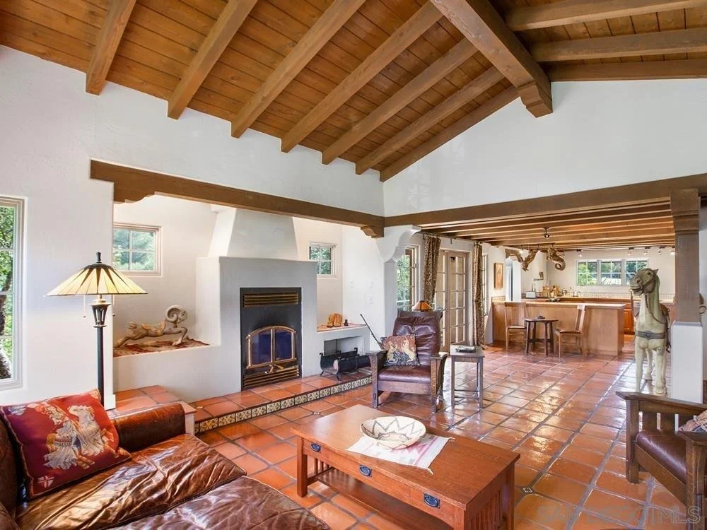Living room with white walls, terracotta tile flooring, wooden ceiling beams, a white fireplace, a leather couch and chair, a wooden coffee table, a floor lamp, and decorative items, with a view into the kitchen area in the background.