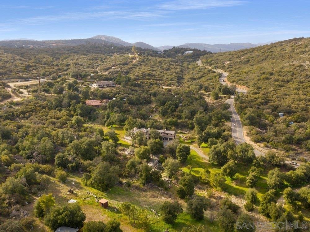 Aerial view of a hilly rural area with winding roads, trees, and scattered houses, under a blue sky.