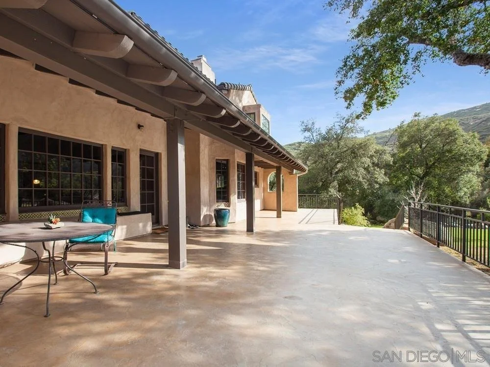 Large outdoor patio with beige stucco house walls, six large windows, brown trim, and a gray roof. There is a metal patio table with a turquoise cushion on one chair, a large ceramic planter, and a small potted plant on the table. Metal railing on th