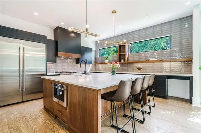 Modern kitchen with a large island, stainless steel refrigerator, black cabinets, white countertop, wooden accents, bar stools, and a wide window with a view of greenery.