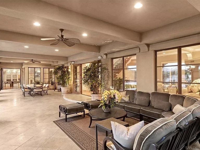 Spacious indoor lounge area with gray sectional sofa, coffee table with flowers, potted plants, glass doors, and ceiling fans.