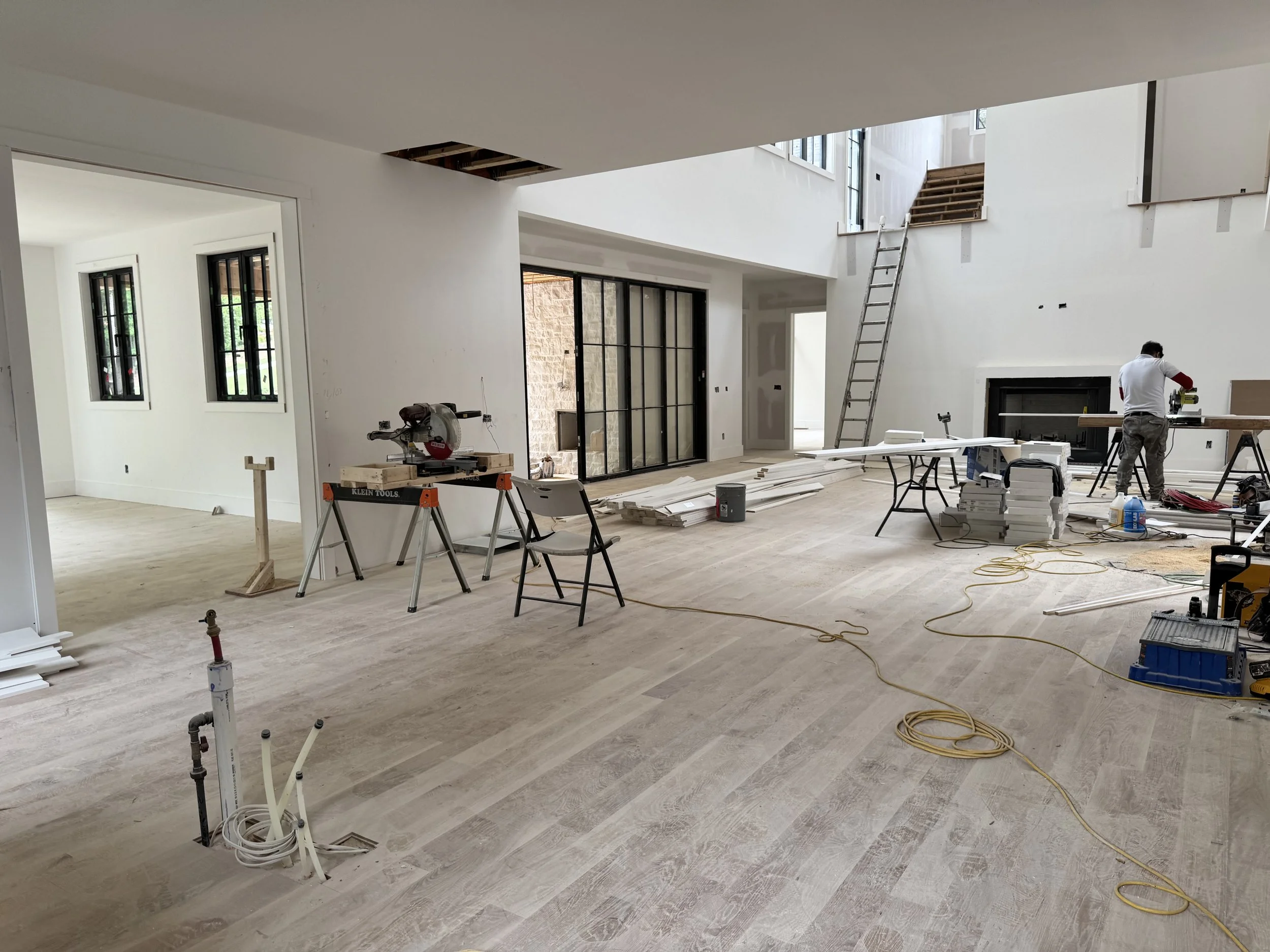 Interior of a house under construction or renovation with unfinished walls, open ceiling panels, construction tools, and a person working on a table.