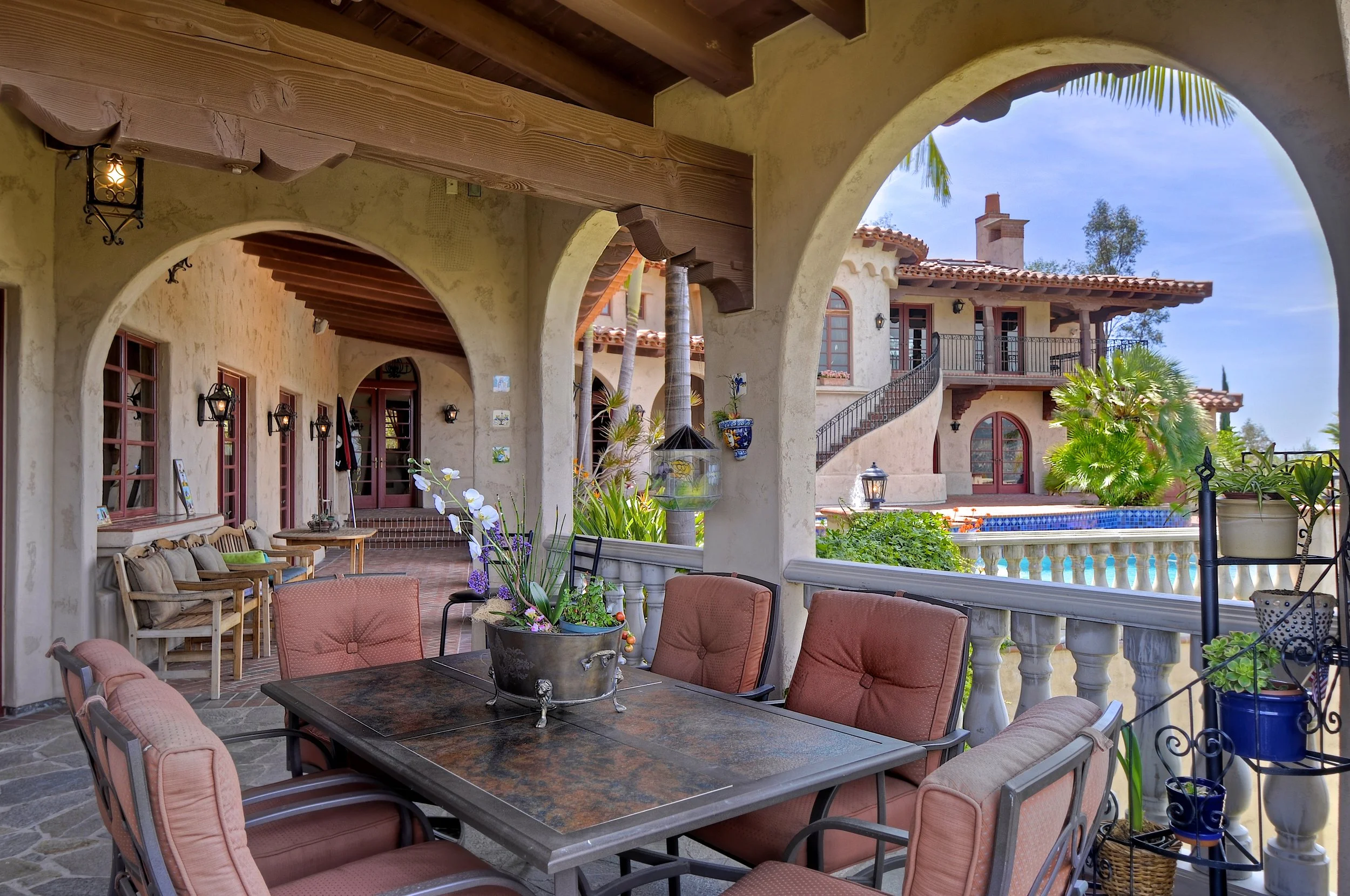 Outdoor patio area with cushioned chairs around a large table, potted plants, arched openings, and a view of a house with a tiled roof, balcony, and lush greenery.