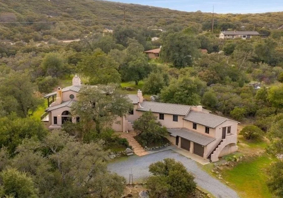 A large house with a beige exterior and tiled roof, surrounded by lush green trees and hills in the background, with a gravel driveway leading up to the garage.